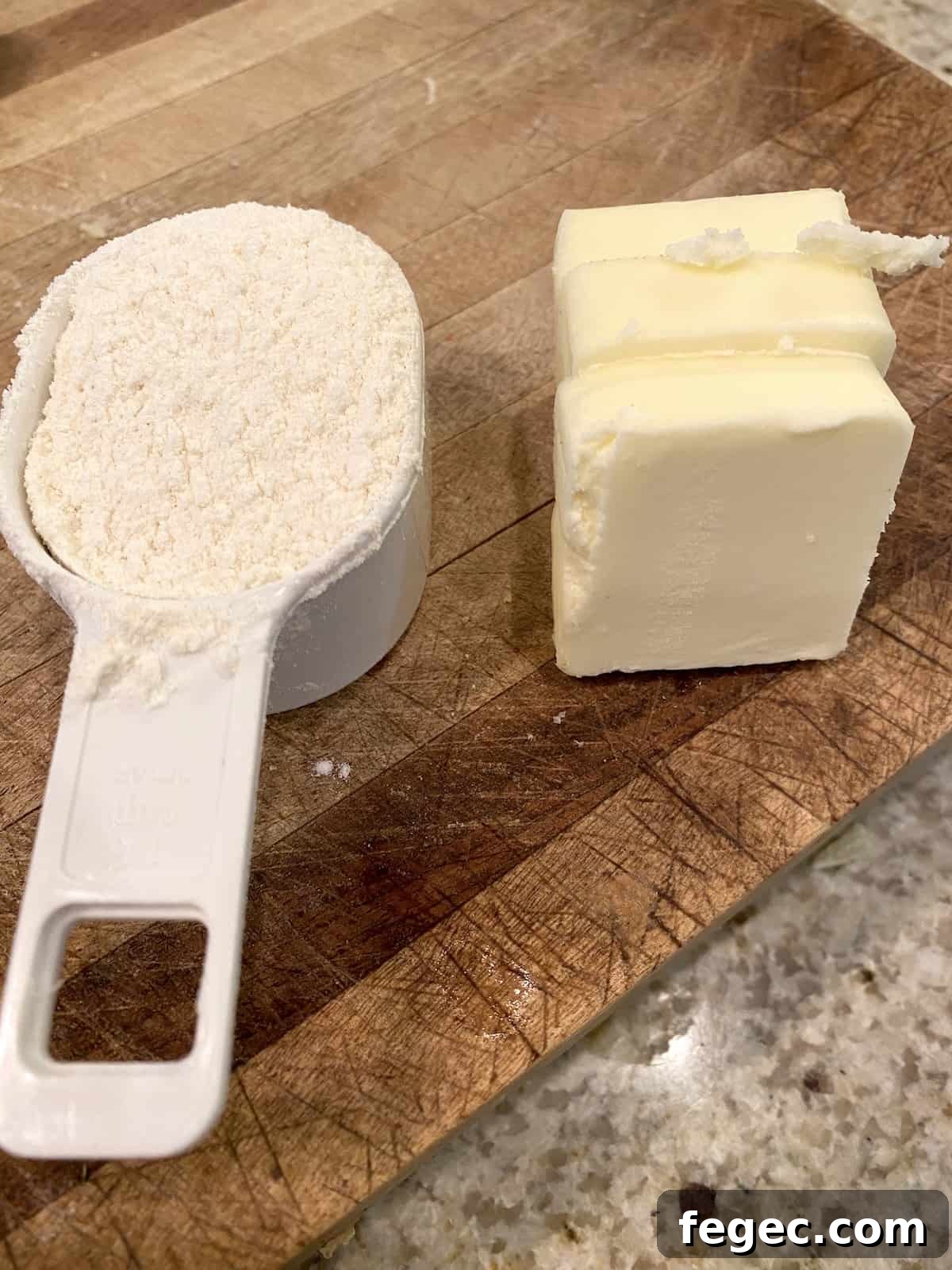 A measuring scoop full of flour and a wedge of butter on a cutting board.