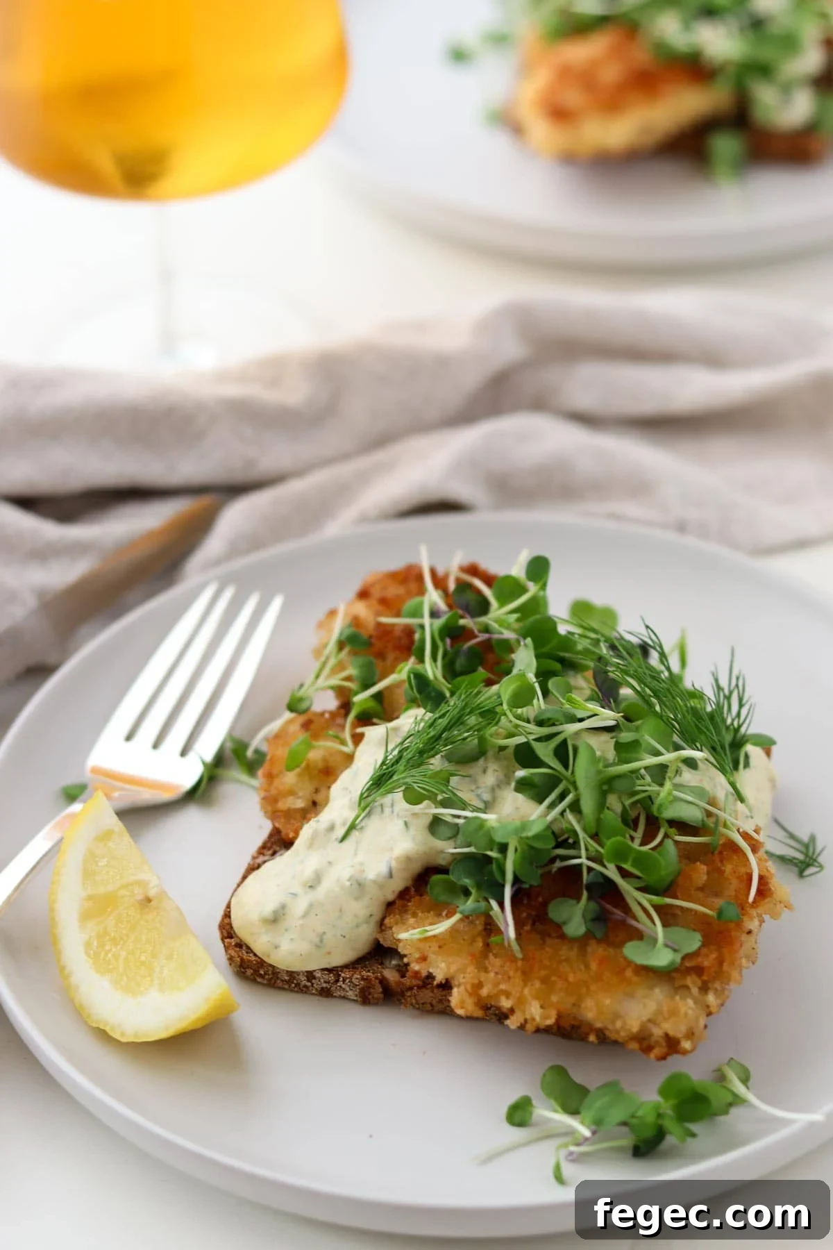 Pan-fried fish on rye topped with sauce and greens with a lemon wedge, fork and beer.