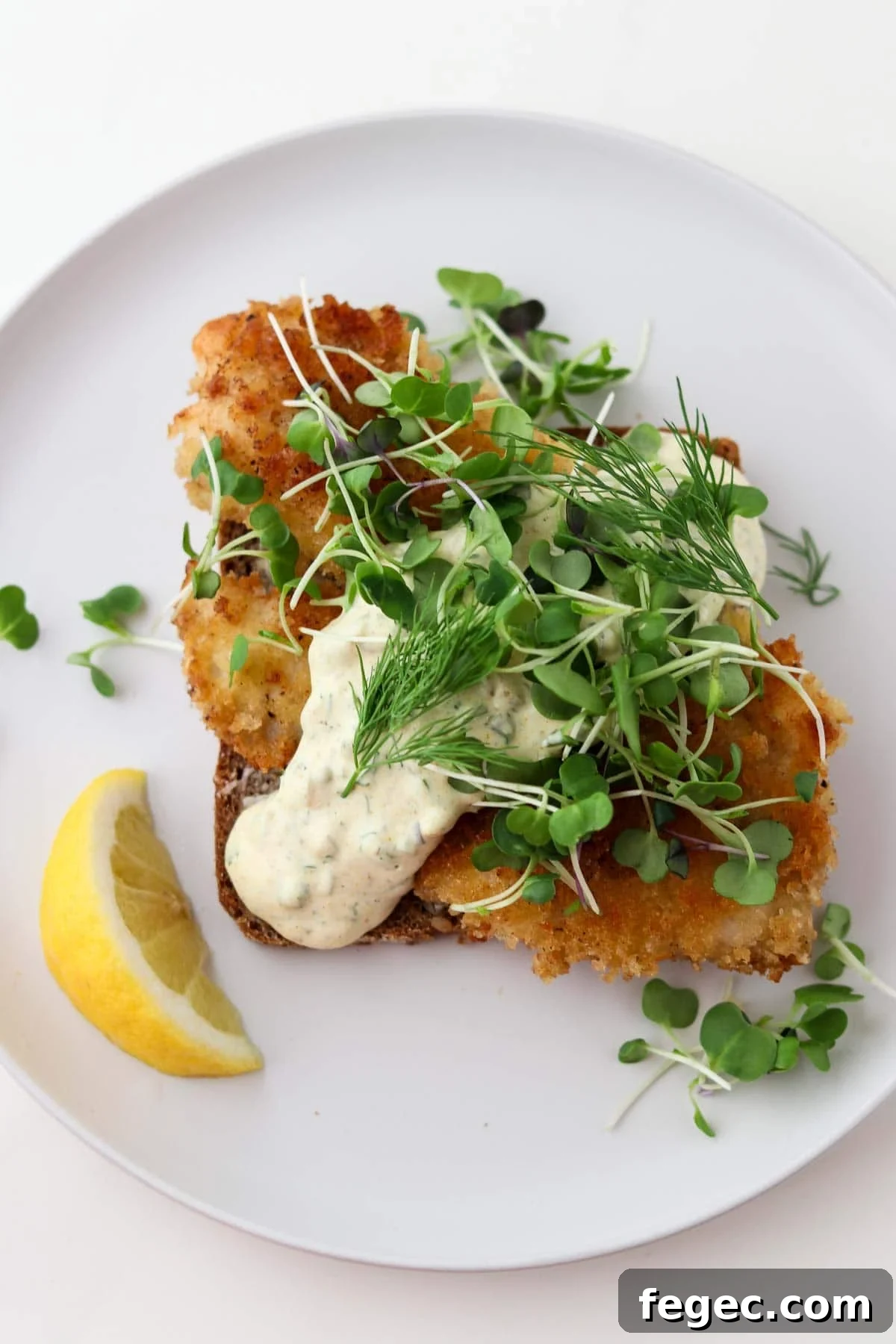 Pan-fried fish on rye bread topped with sauce, greens and with a lemon wedge.