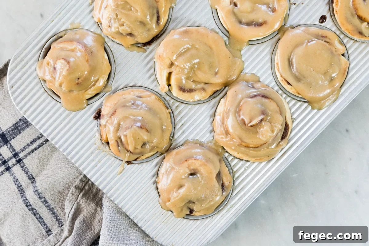 Top down view of silver muffin in containing frosted cinnamon roll muffins with towel on top of marble surface