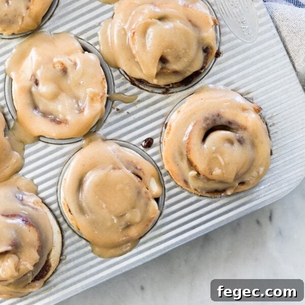 Top down view of silver muffin in containing frosted cinnamon roll muffins with towel on top of marble surface