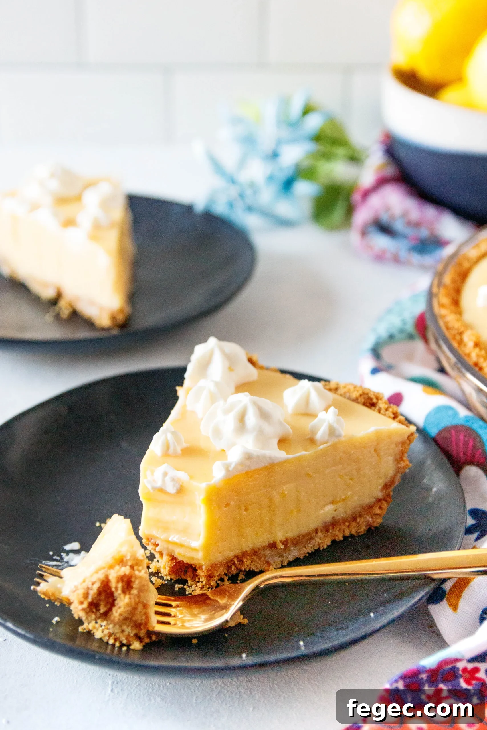 angled picture of the creamy lemon pie slice on a black plate with a gold fork holding a piece. There's another slice in the back left of the picture and a portion of the whole pie in the back right