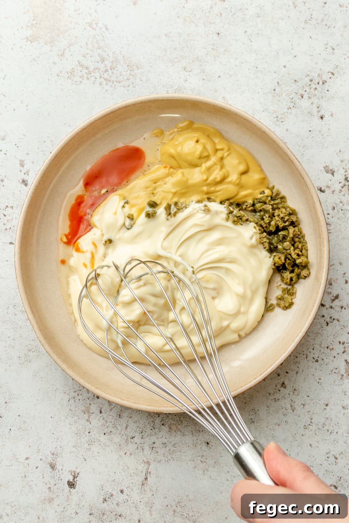A remoulade sauce is whisked in a ceramic shallow bowl on a light grey surface.
