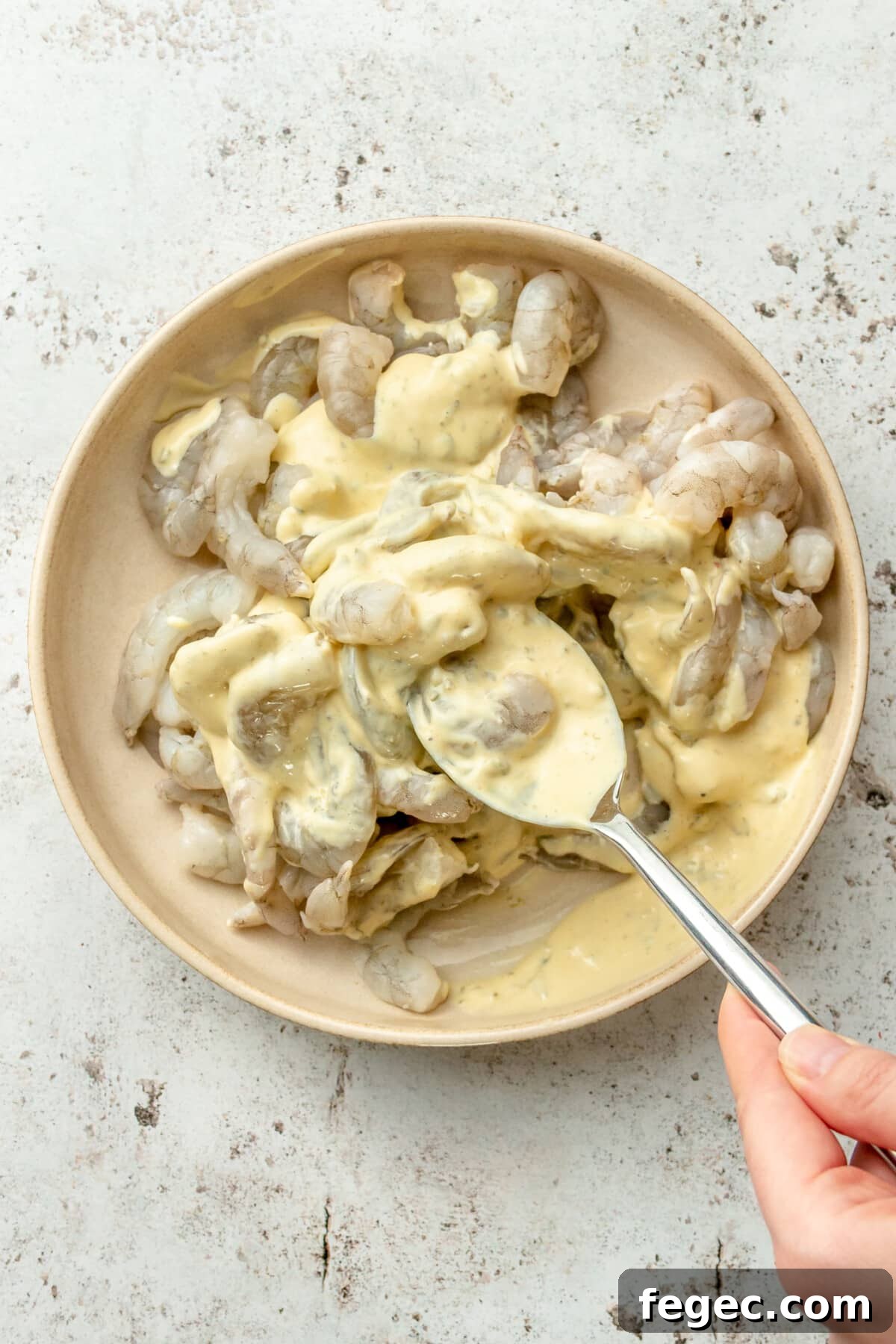 Shrimps are tossed through a remoulade sauce in a shallow ceramic bowl on a light grey surface.