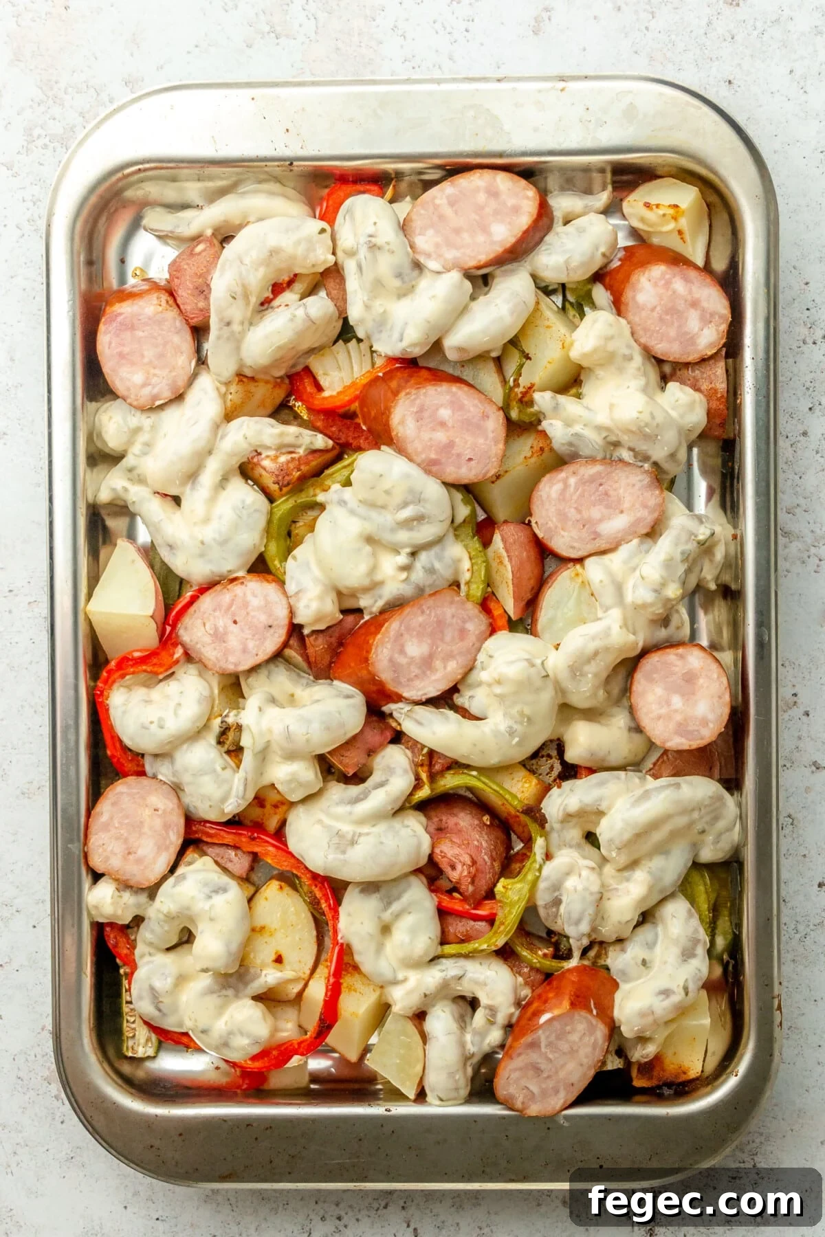 Shrimp coated in a remoulade sauce and andouille sausage pieces sit on top of roasted vegetables on a rimmed stainless steel baking sheet on a light grey surface.
