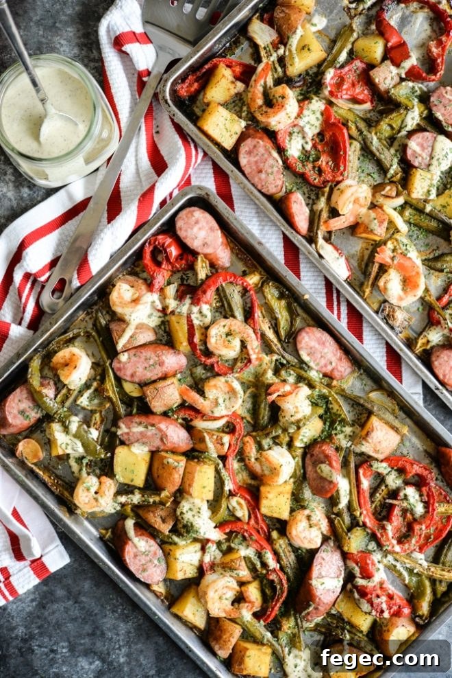 overhead view of a sheet pan loaded with sausage, shrimp, potatoes, asparagus, and red bell peppers with a creamy sauce drizzled over it on top of a dark grey table and red striped napkin