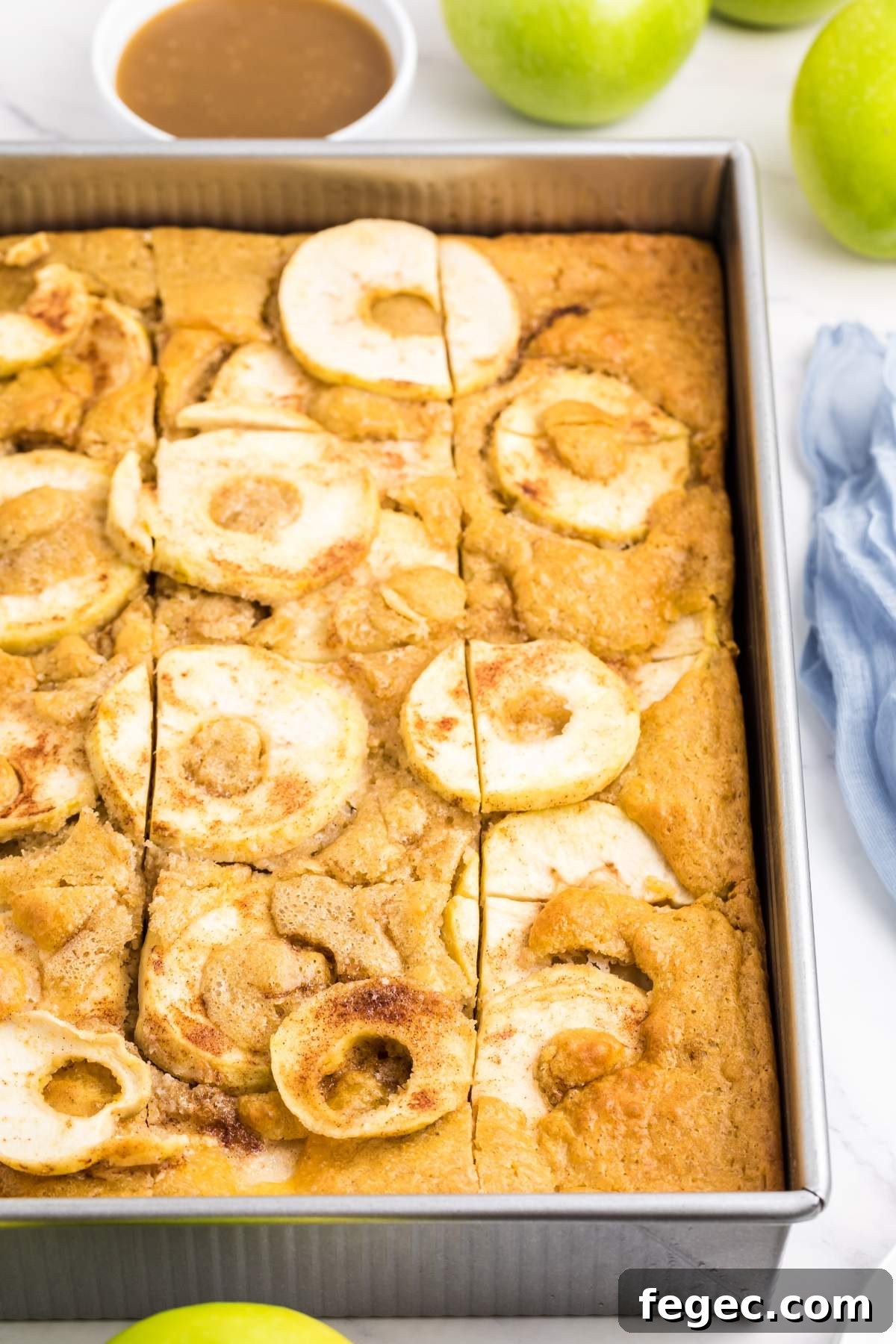 A pan of apple slab cake cut into squares, topped with apple slices, with green apples and a cup of sauce in the background.