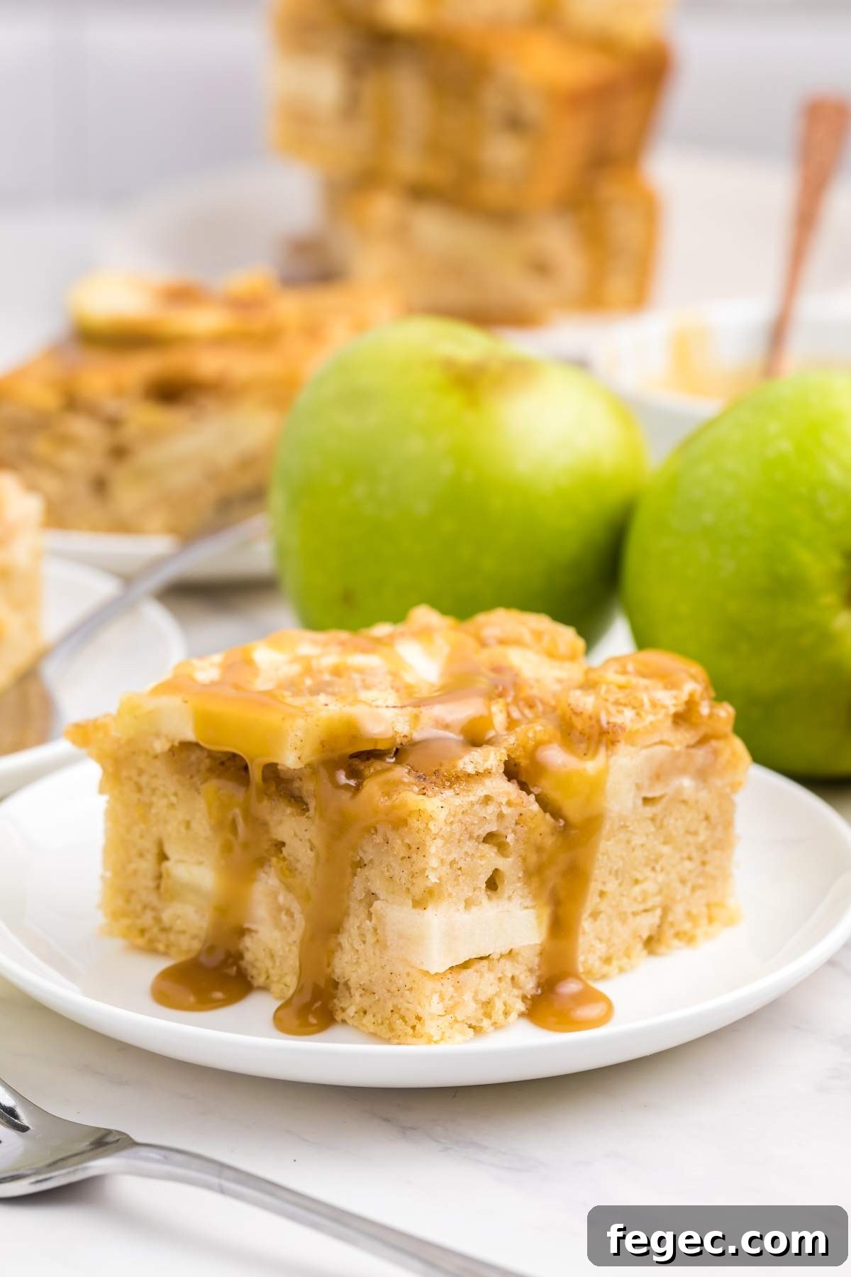 A slice of apple sheet cake topped with caramel sauce on a white plate, with two green apples in the background.