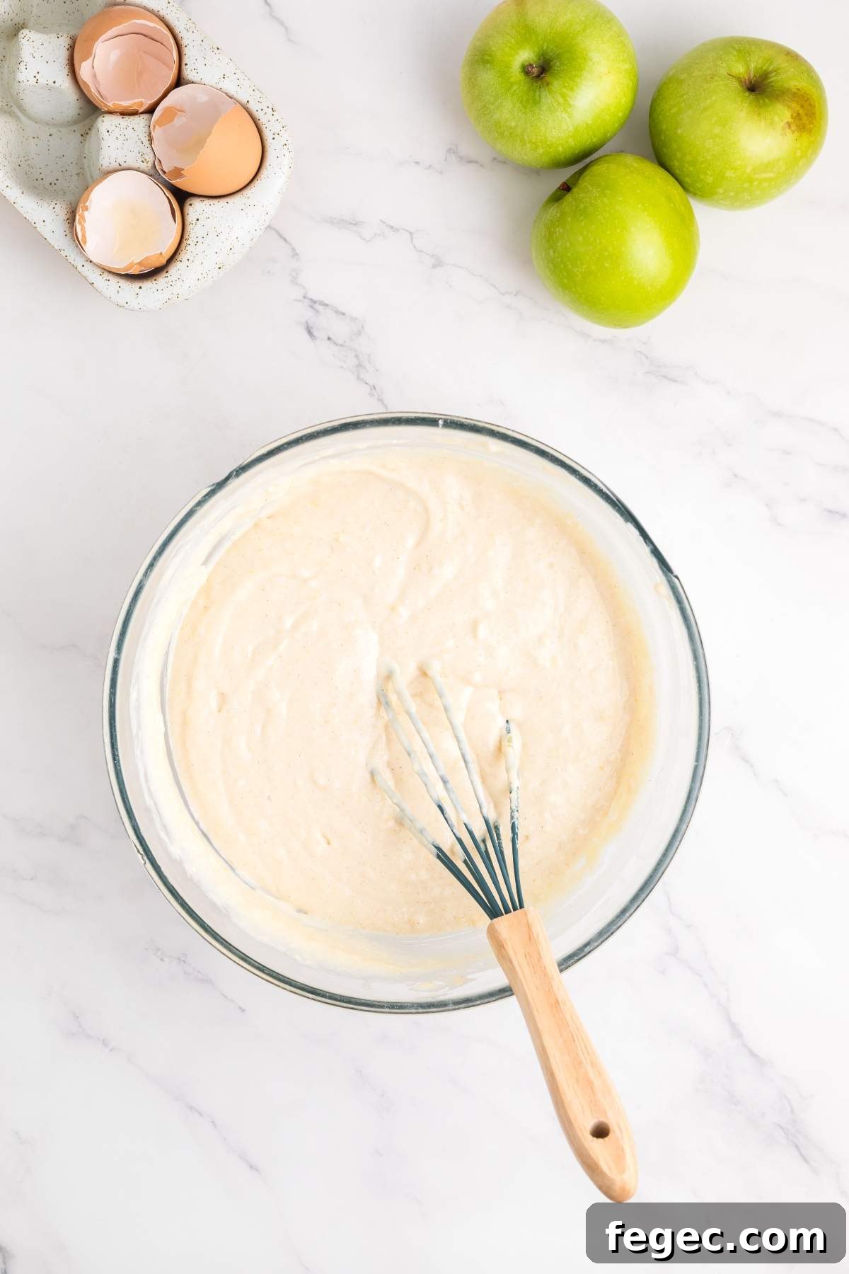 A mixing bowl filled with batter and a whisk rests on a counter alongside three green apples, hinting at an apple sheet cake in the making, with a cracked egg shell in a container nearby.