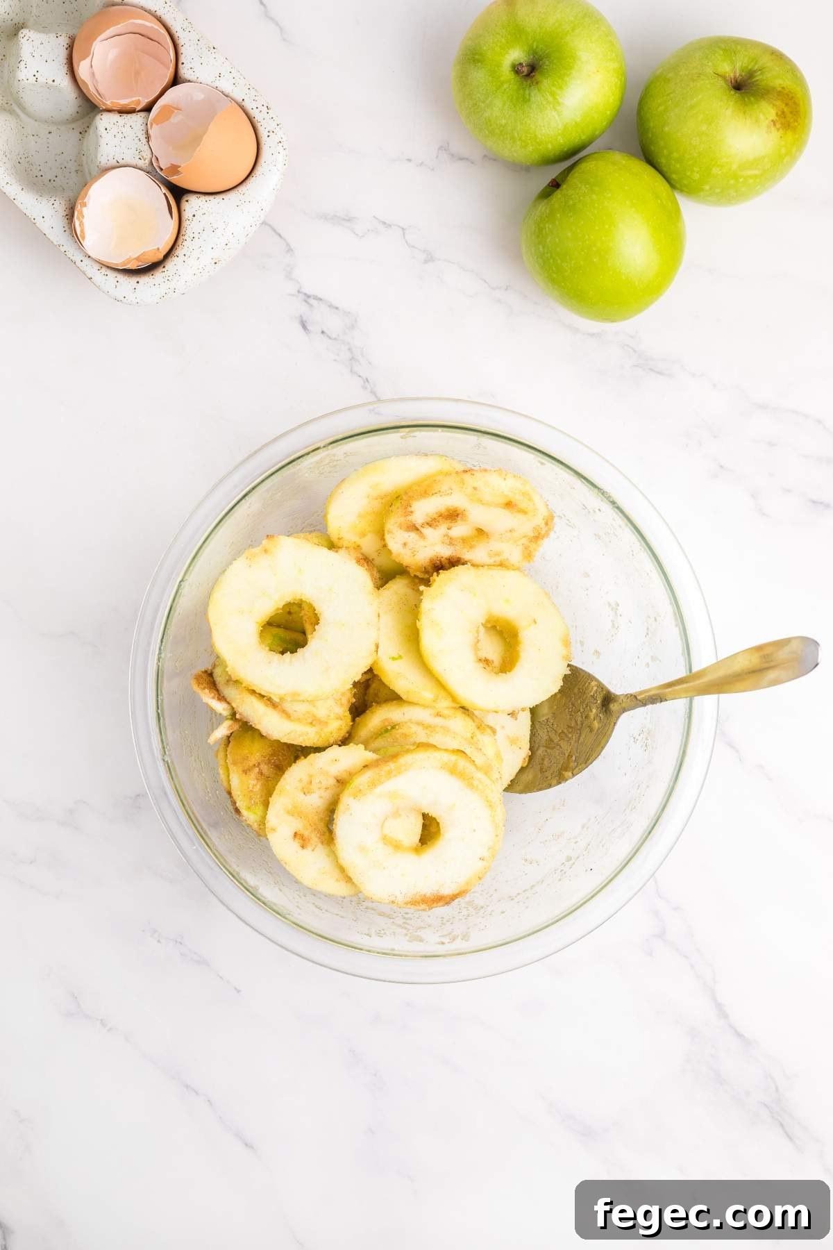 A glass bowl with peeled and cored apple slices and a spoon. Beside it, there are three green apples and a container holding cracked eggshells, perfect ingredients for that apple sheet cake you're planning to bake.