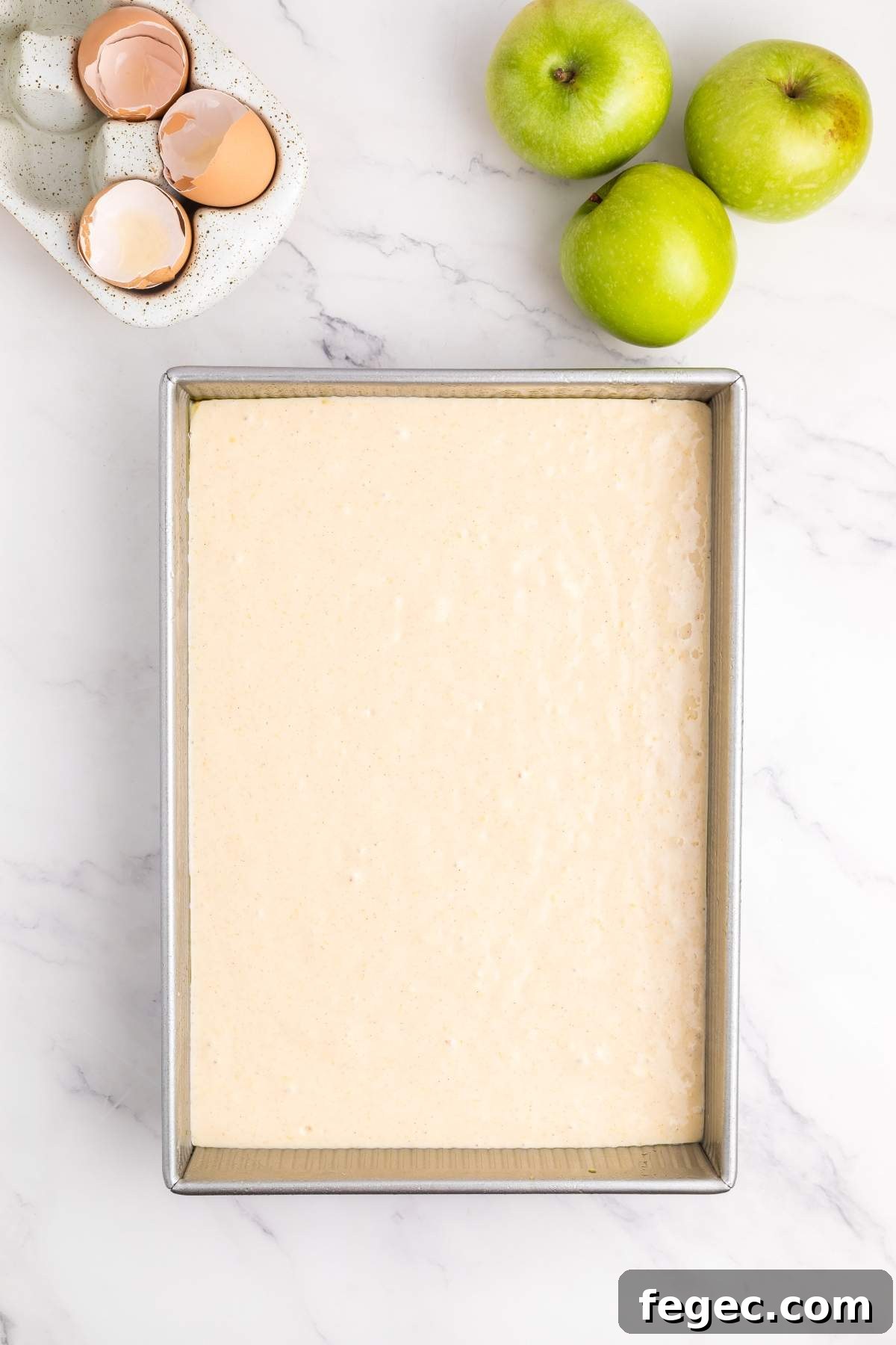 A baking pan filled with apple sheet cake batter is placed on a marble surface, next to three green apples and a bowl with cracked eggshells.