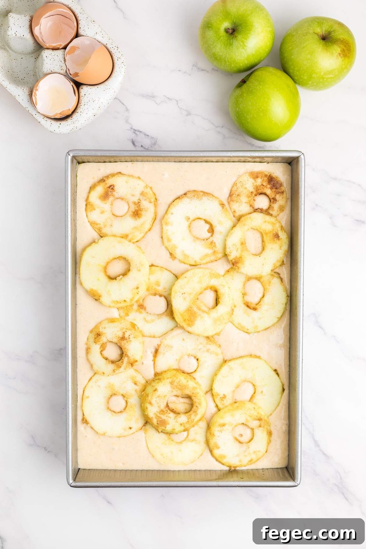 A baking pan filled with a mixture for an apple sheet cake, topped with round apple slices. Beside the pan, three green apples and two cracked eggshells in a dish rest on a white marble surface.