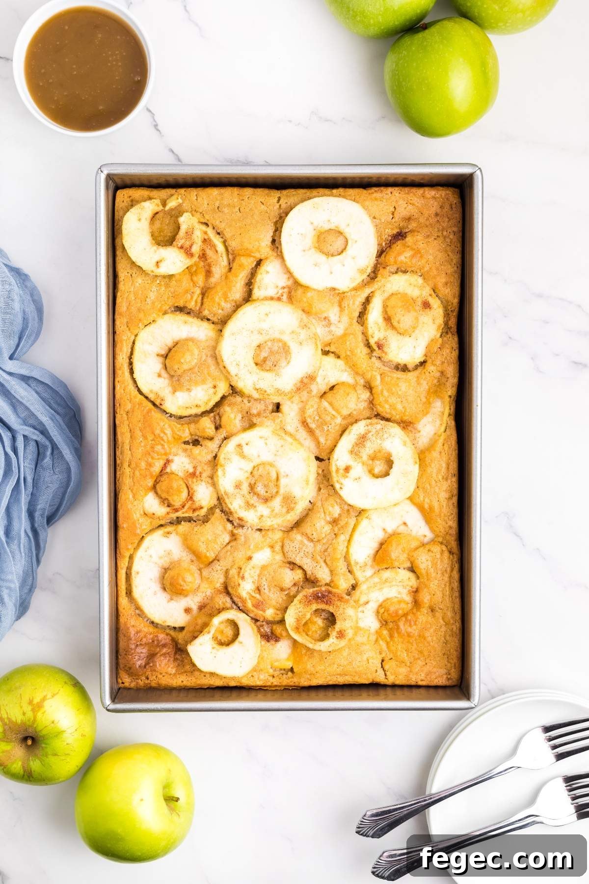 A baked apple sheet cake in a rectangular pan, topped with apple slices. Surrounding the pan are green apples, a blue cloth, a small cup with sauce, forks, and a white plate on a white marble surface.