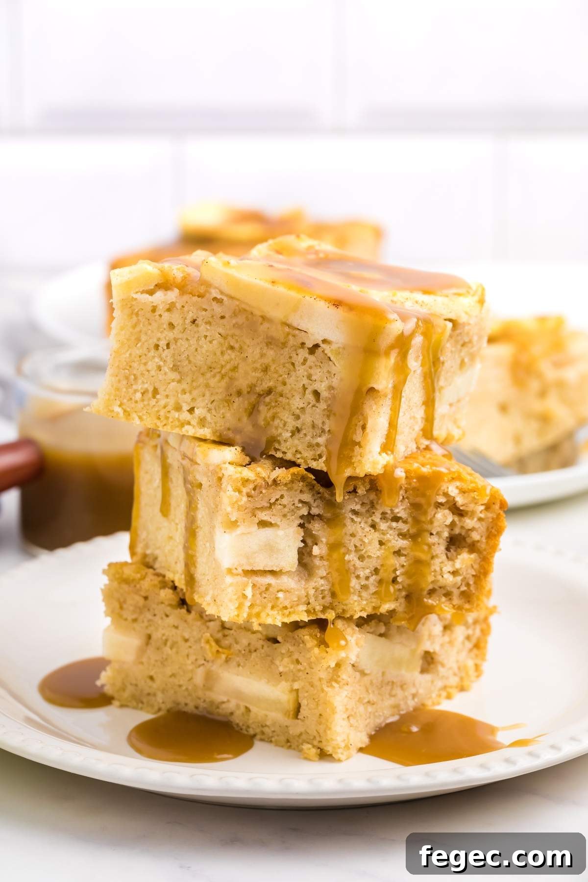 A stack of caramel-drizzled apple blondies on a white plate, resembling the coziness of an apple sheet cake, with more blondies and caramel in the background.