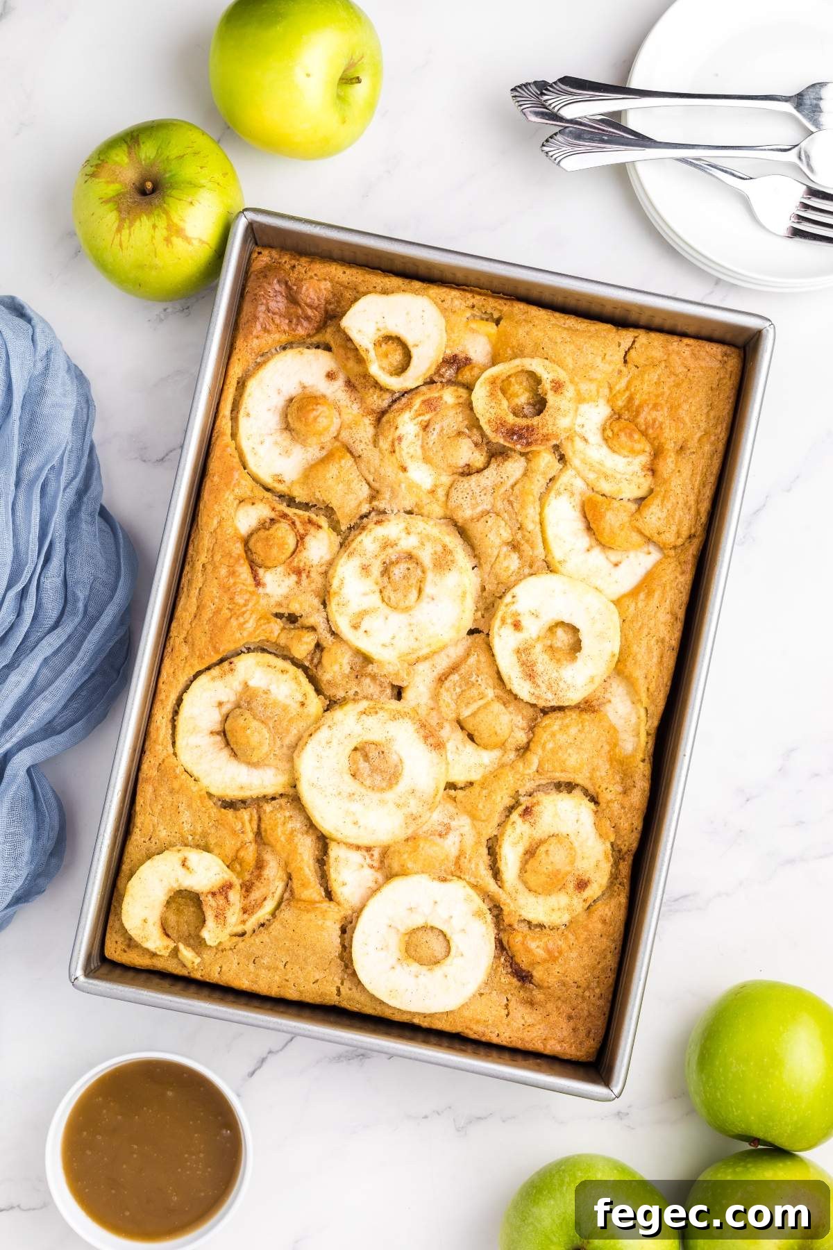 A rectangular apple sheet cake in a baking pan topped with sliced apples. Surrounding the pan are green apples, a small bowl of sauce, a blue cloth, and stacked plates with forks on a marble surface.
