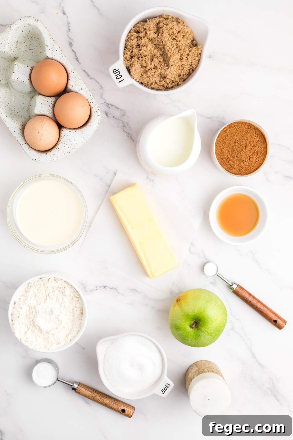 Top view of baking ingredients for an apple sheet cake, including eggs, brown sugar, milk, cinnamon, vanilla extract, flour, butter, an apple, white sugar, salt, and baking powder on a marble surface.