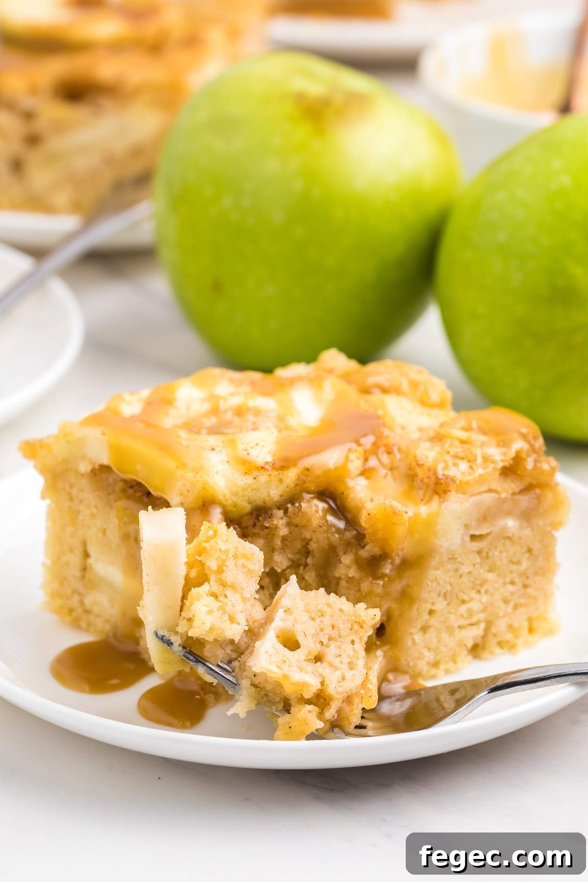 A slice of caramel apple slab cake rests invitingly on a white plate with a fork. Two green apples are positioned in the background, adding a fresh touch.