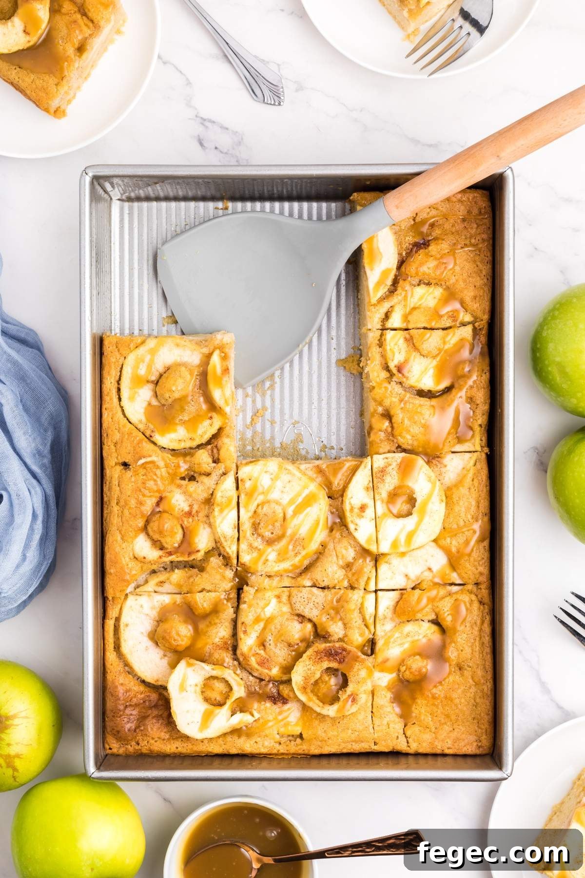 A baking tray with an apple pan cake, one slice missing. Forks, apples, and a dish with caramel sauce surround the tray on a white background. A spatula rests on the tray's edge.