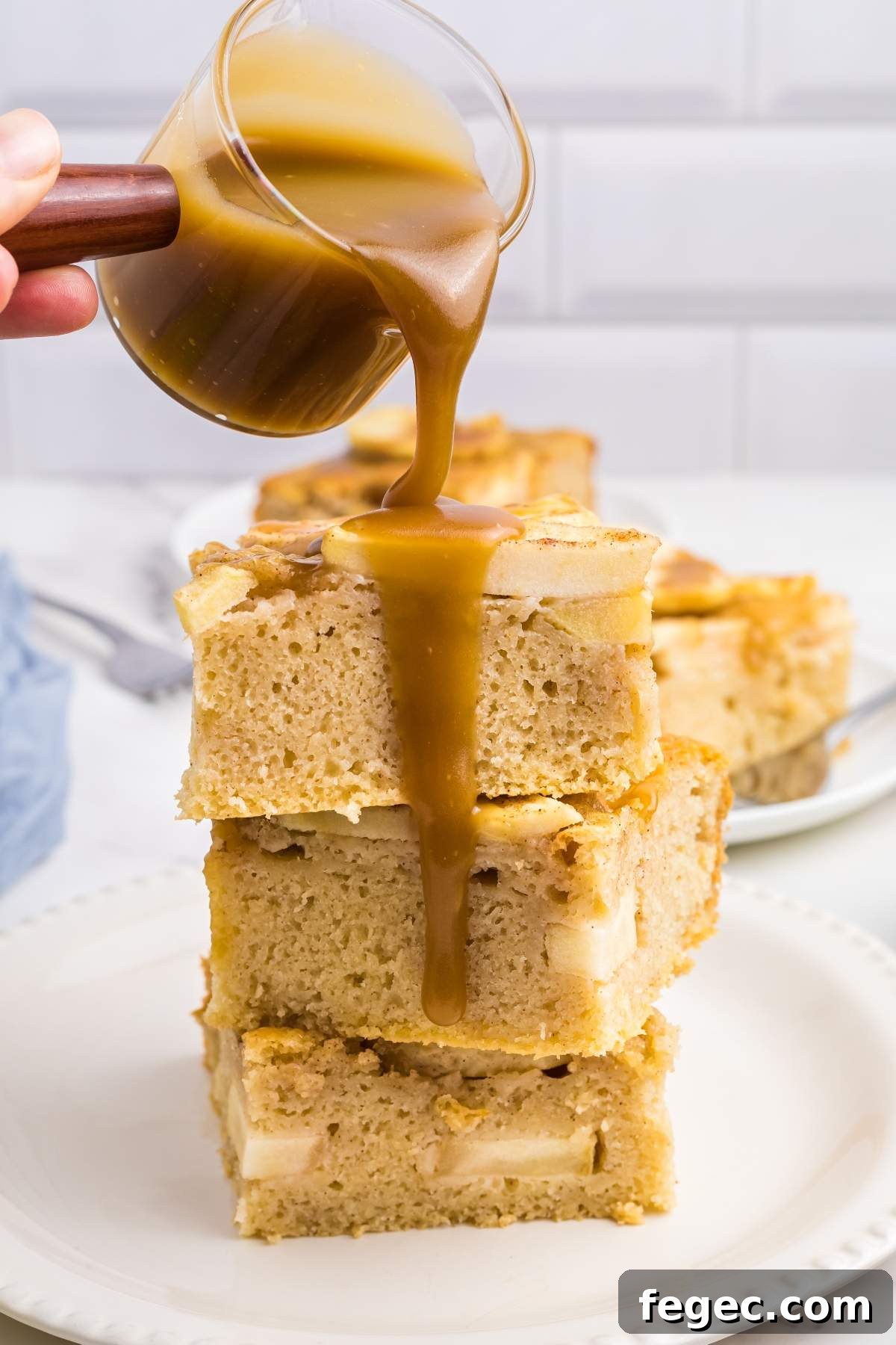 A hand pouring caramel sauce over a stack of three apple sheet cake slices on a white plate, reminiscent of the rich flavors found in an indulgent apple sheet cake.