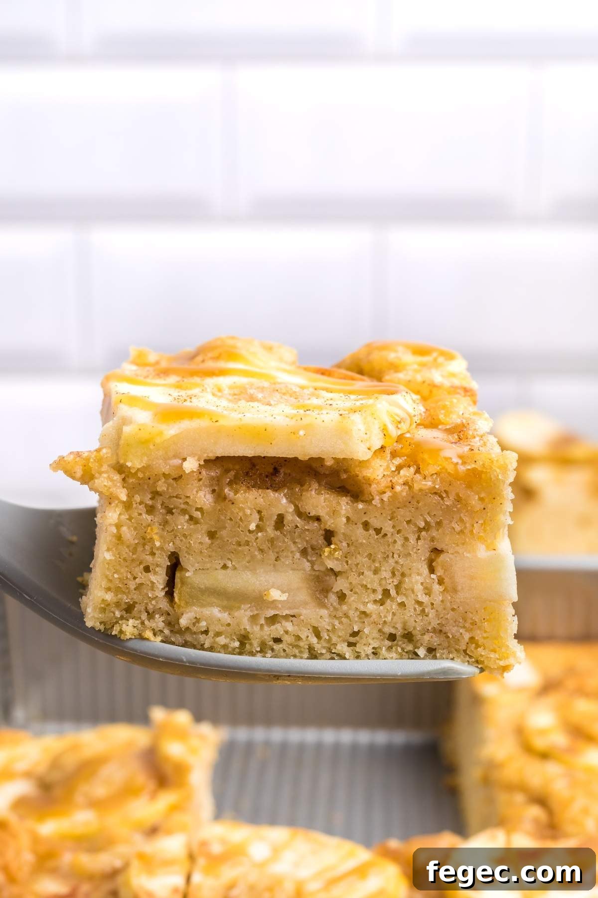 A spatula holds up a piece of apple sheet cake with a golden-brown top, as part of a freshly baked batch seen in a pan in the background.