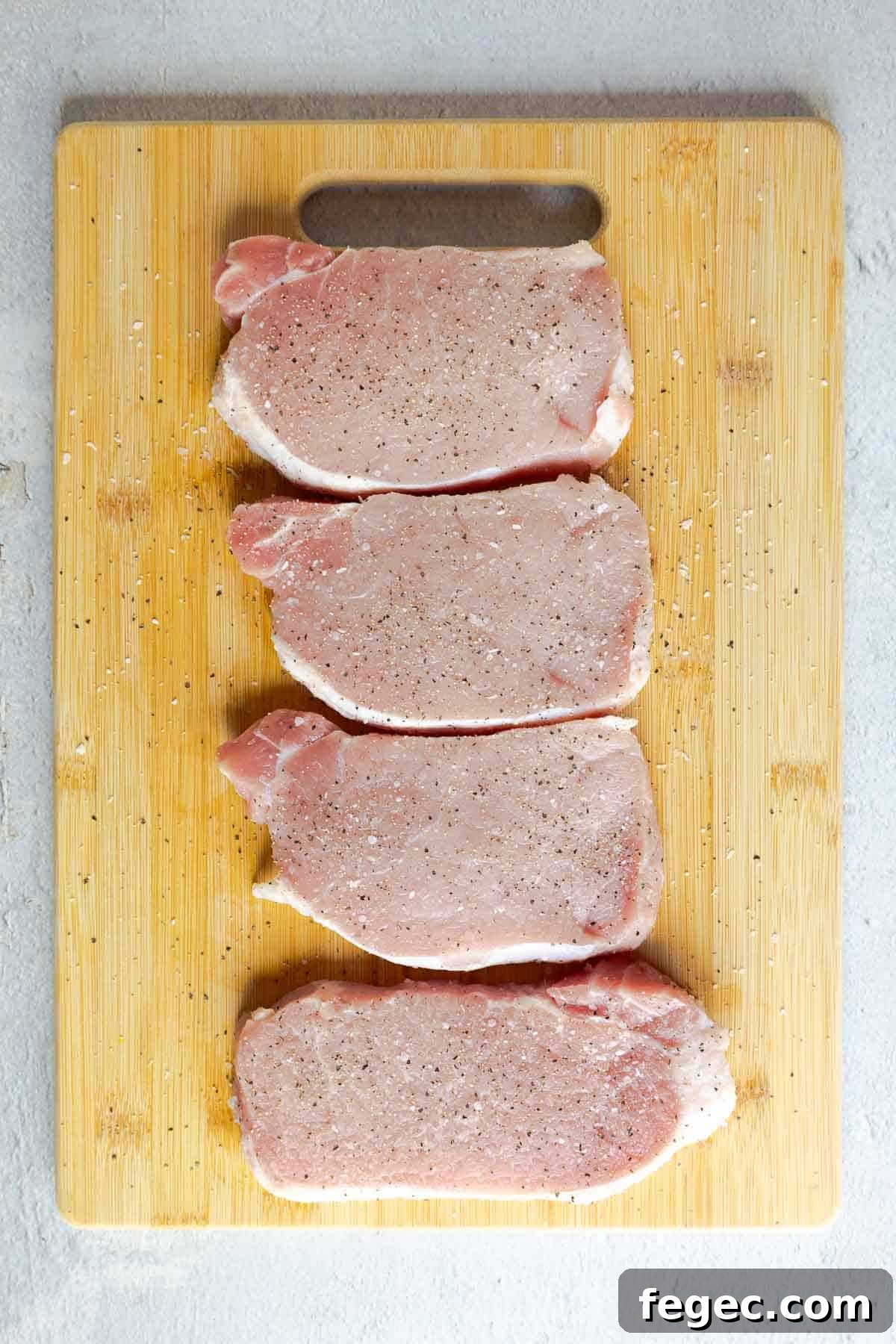 French Onion Pork Chops with Melted Gruyère and Caramelized Onions 5 Four raw pork chops seasoned with salt and pepper on a wooden cutting board.