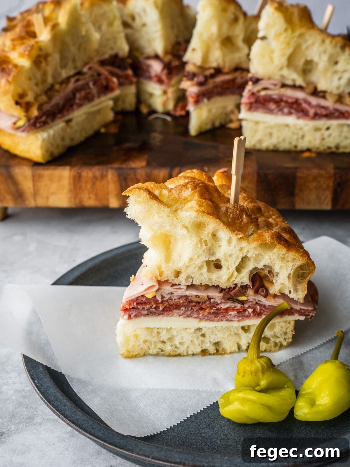 Muffuletta sandwich made with fresh homemade focaccia stuffed with cheese and deli meat on a plate with a side of hot peppers.