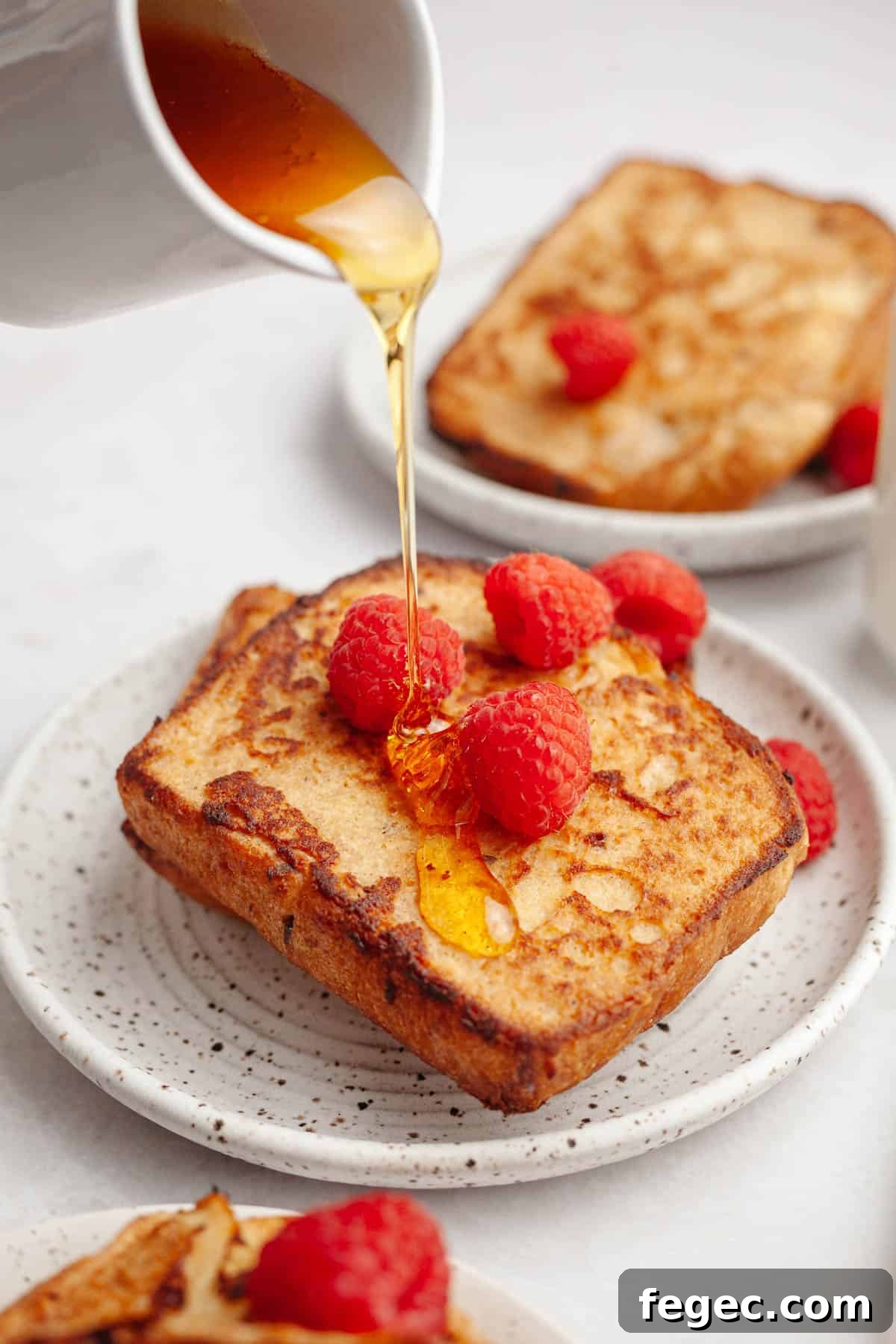 Two slices of homemade buttermilk french toast on a small plate, topped with raspberries and maple syrup being poured on top.