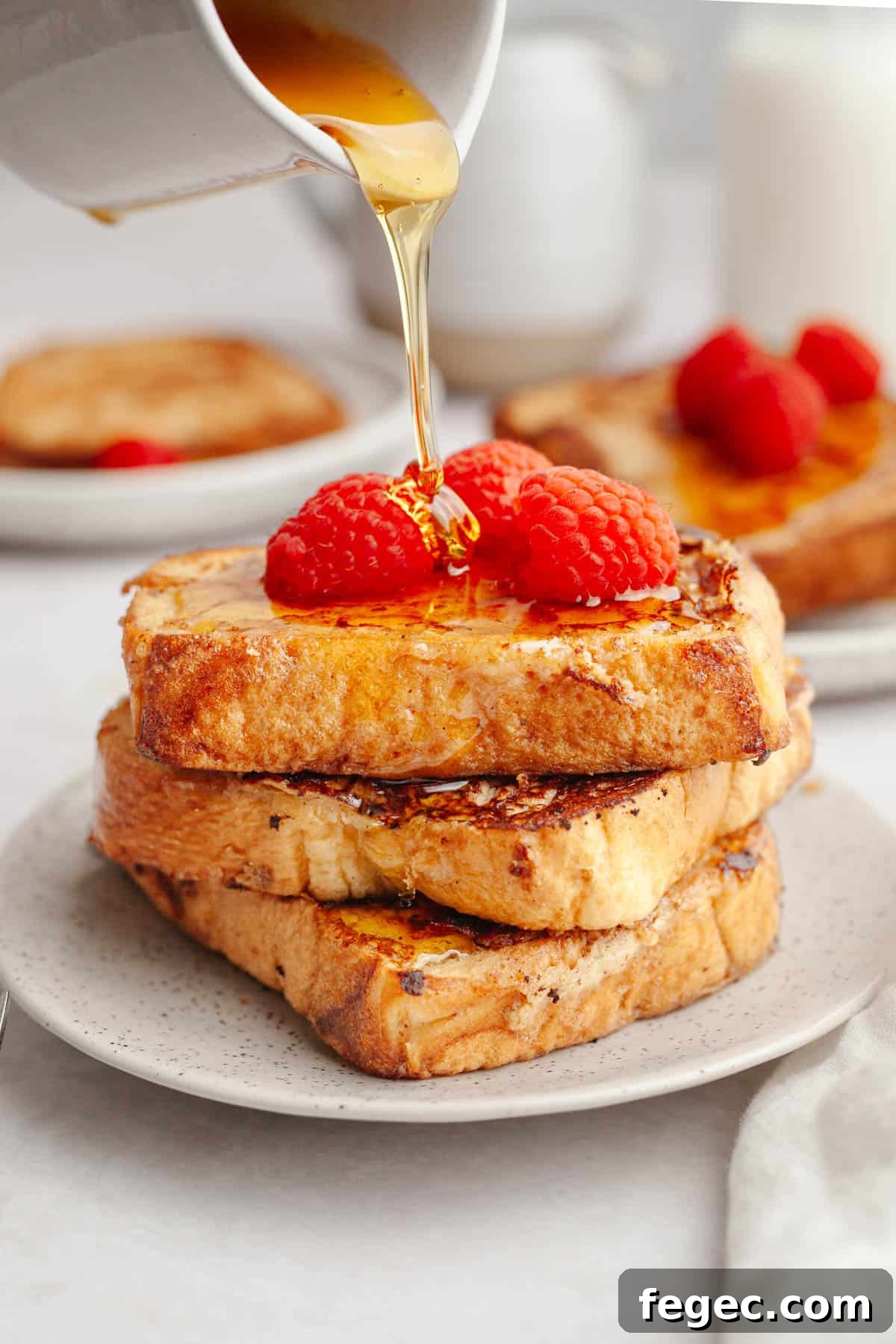Three slices of homemade buttermilk french toast on a small plate, topped with raspberries and maple syrup being poured on top.