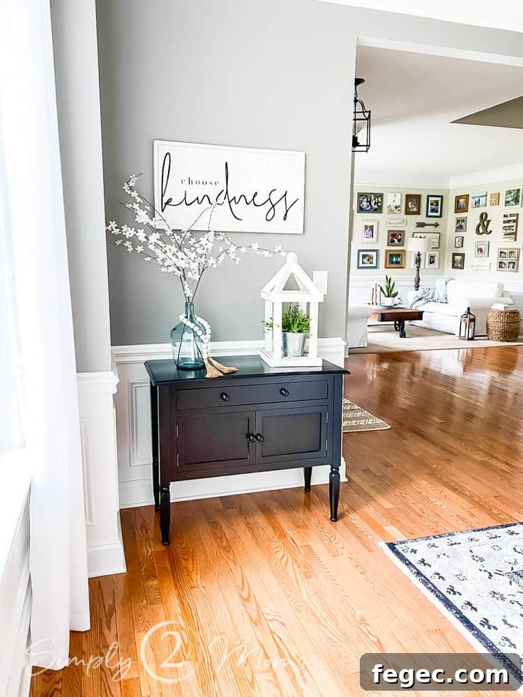 Spring Dining Room Ideas for Greenery Lovers 11 small side table with 2 drawers and double cabinet below. choose kindness word artwork hung on wall above table. glass jar with white floral stems and wood bead around jar and wood lantern with faux plant inside on top of table. living room beyond dining room can be seen in photo