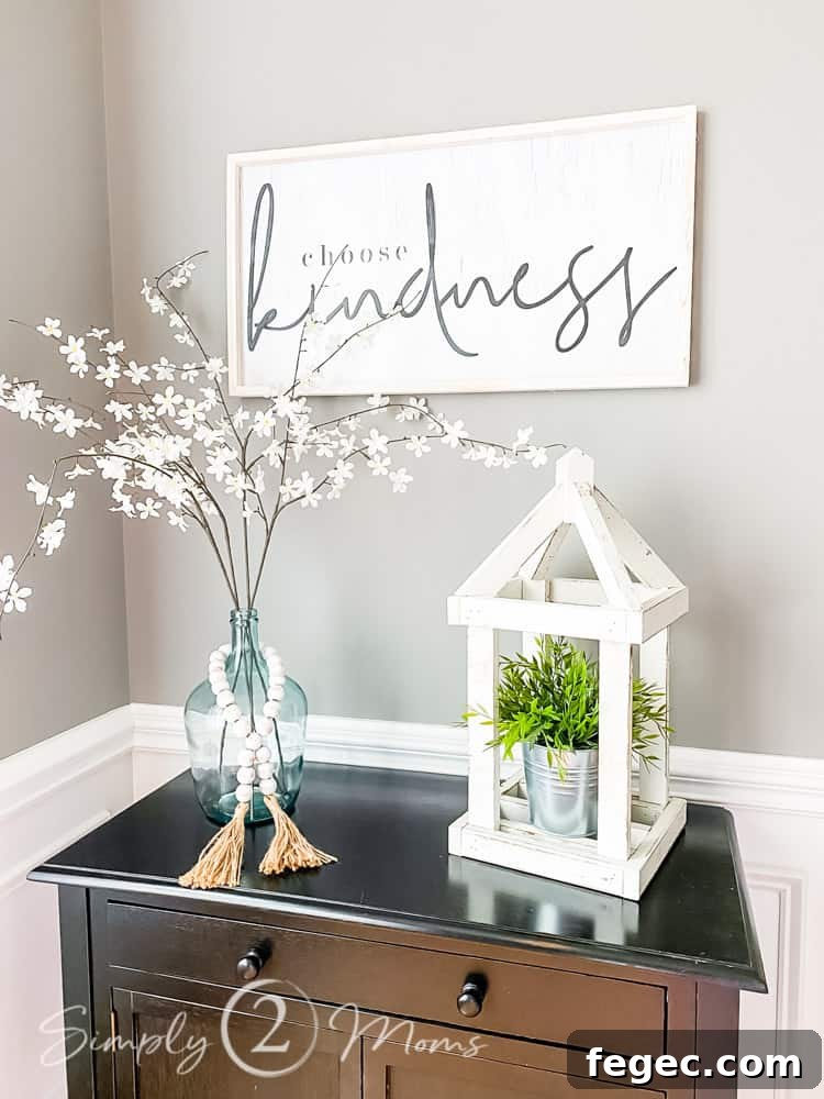 Spring Dining Room Ideas for Greenery Lovers 12 close up of table top with glass bottle neck jar filled with faux floral stems and string of beads around jar wood lantern next to jar with faux plant inside and word art hung on wall above table