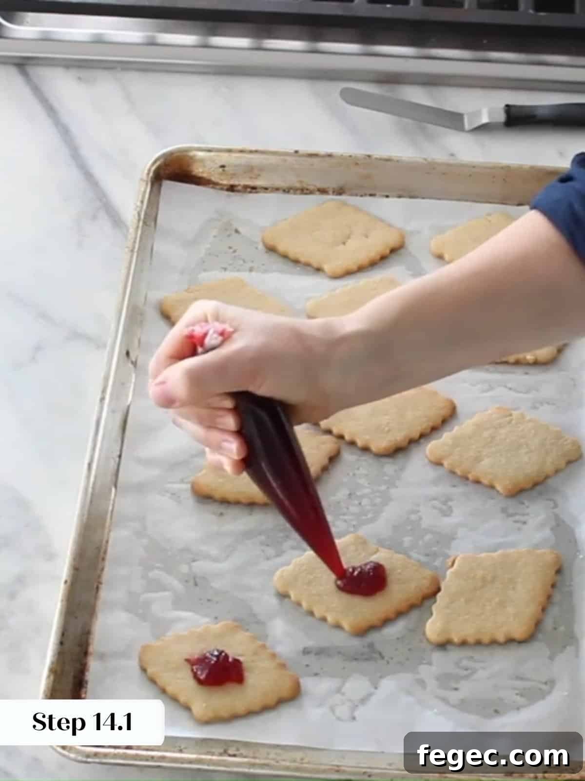 Raspberry preserves being piped onto the flat side of cookie bottoms before sandwiching.
