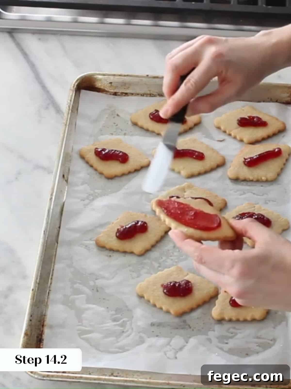 Raspberry jam being spread over baked cookie bottoms before adding the sugar-dusted tops.