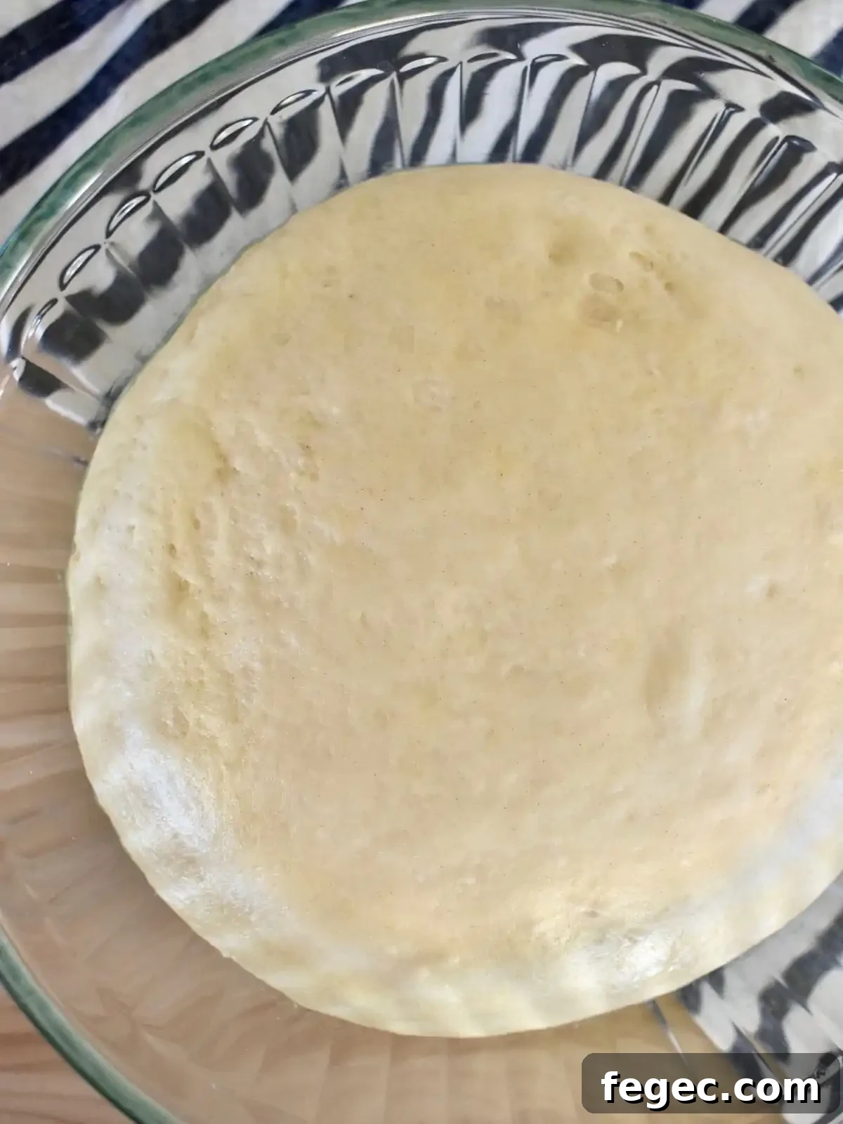 Pizza dough risen in a glass bowl on top of a striped napkin.