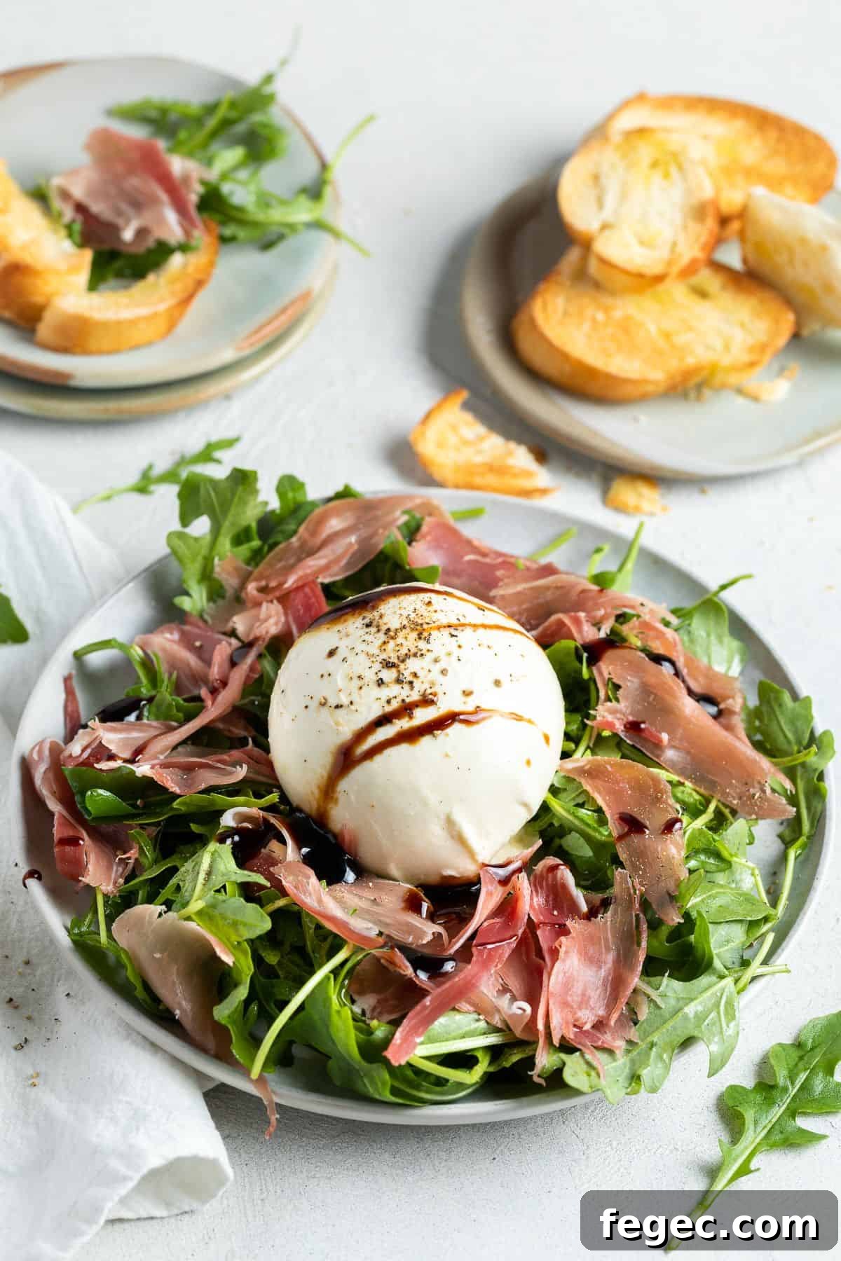 Burrata and prosciutto appetiser sitting on a round plate, with a dish of toasted bread in the corner.