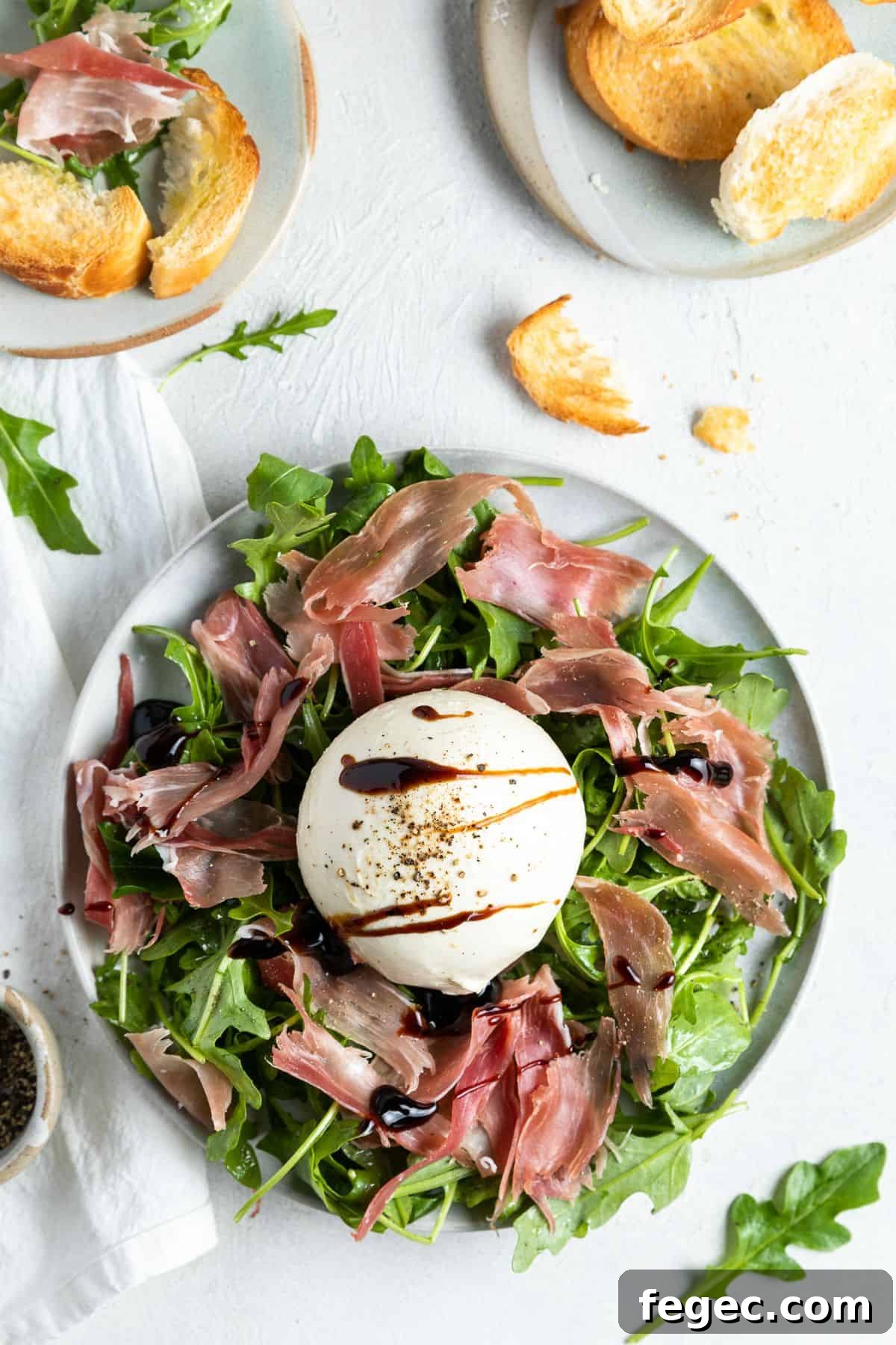 Burrata and prosciutto appetiser sitting on a round plate, with a dish of toasted bread in the corner.