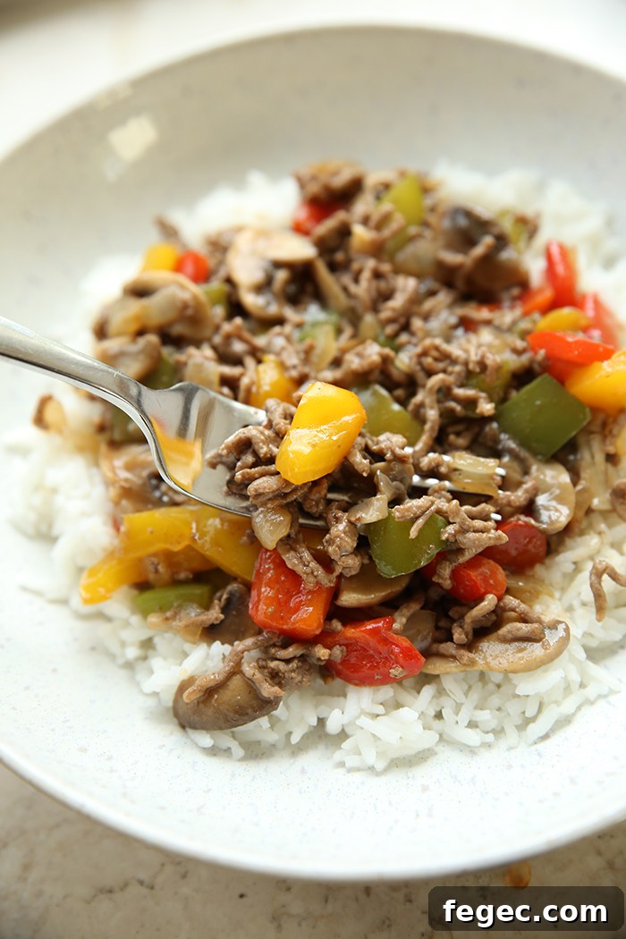 Diced bell peppers and onions, sliced mushrooms, and ground beef in a brown gravy served over white rice in a curved dinner plate. 