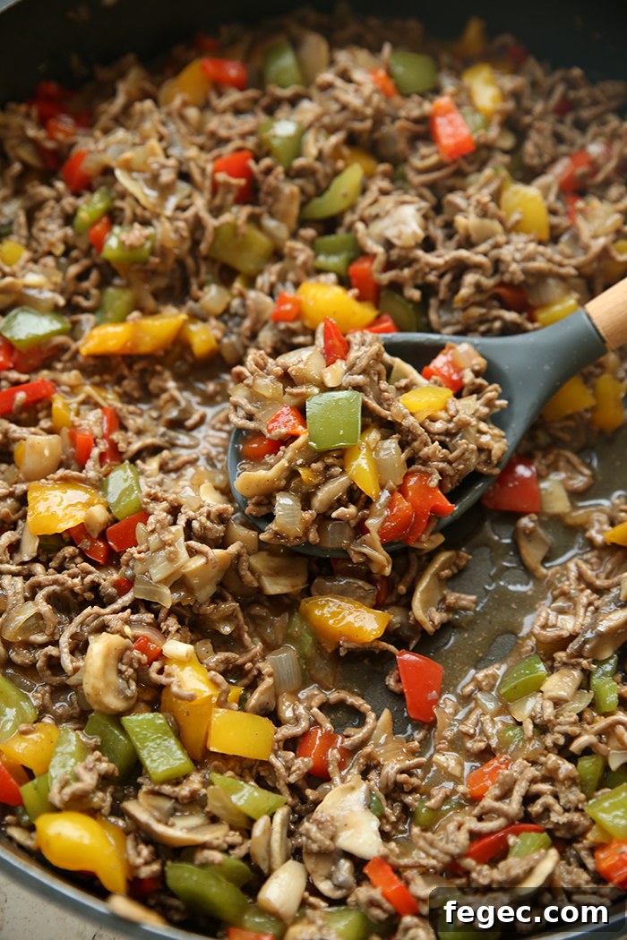 A large skillet of ground beef, bell peppers, onions and mushrooms in a brown gravy sauce. A serving spoonful is held on top of the mixture from the right.