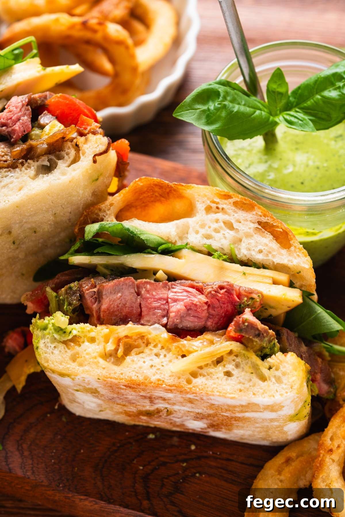 Close-up of a steak sandwich with cheese, spinach, and sauce, with onion rings and green dipping sauce nearby.