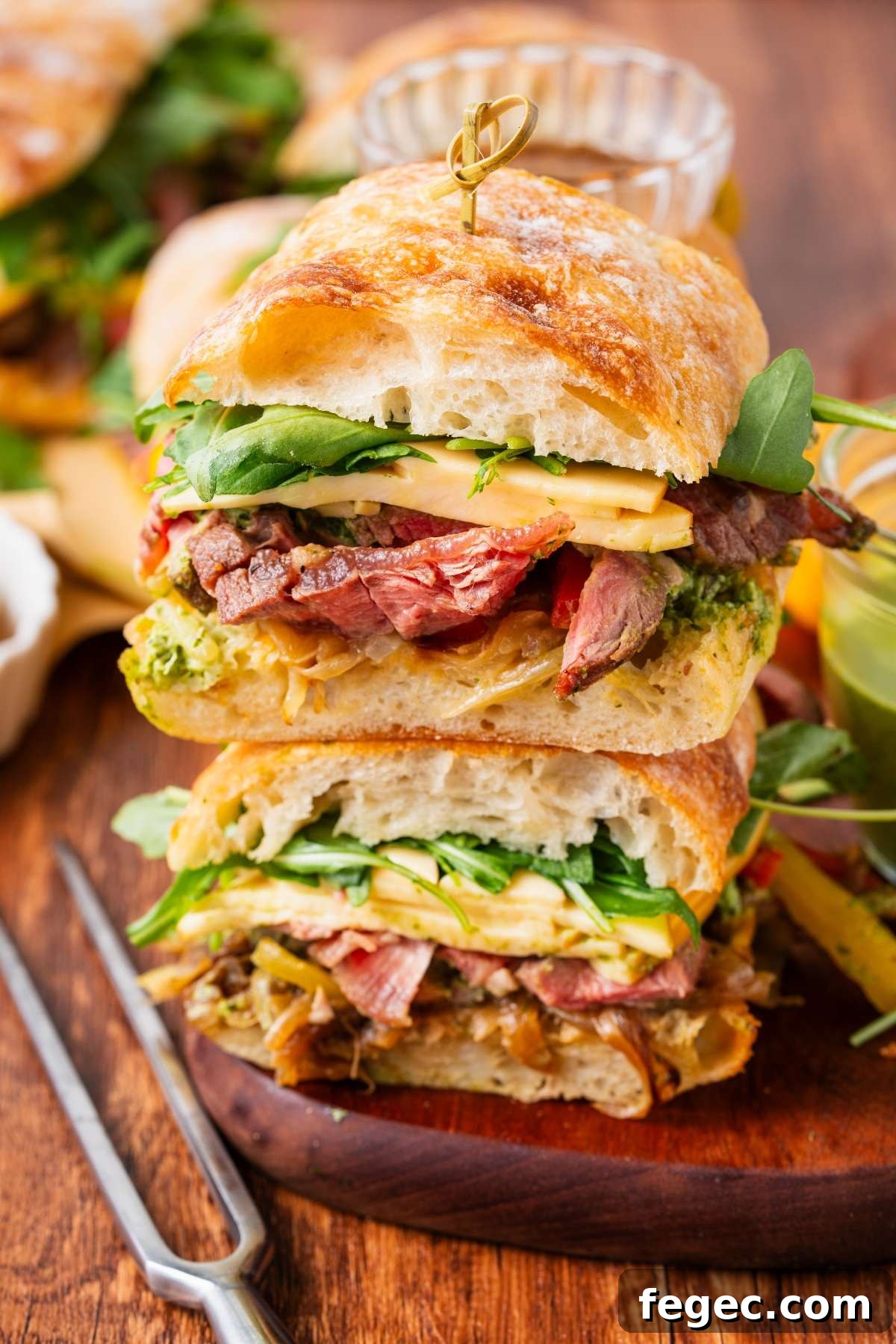 Two stacked steak sandwiches with greens, cheese, and ciabatta bread on a wooden surface.