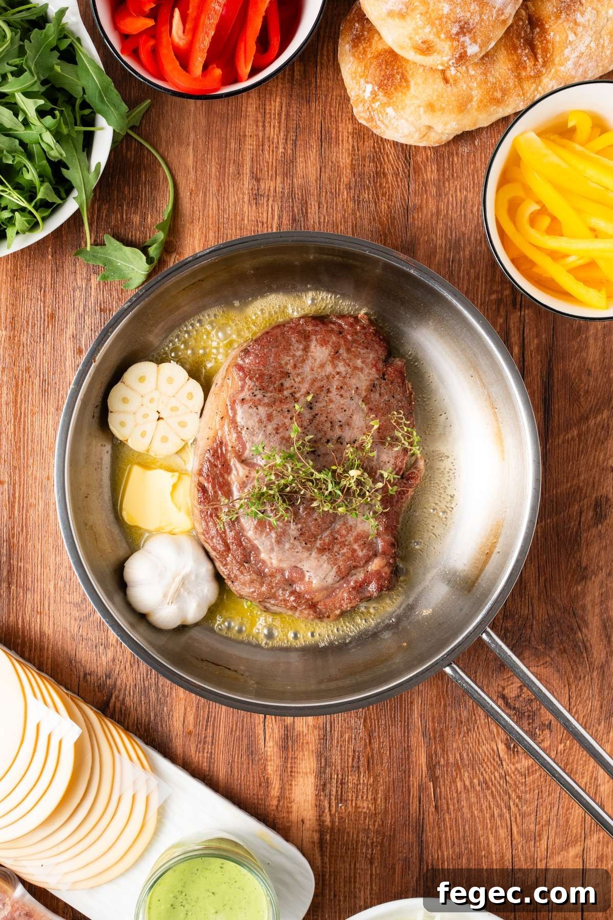 A steak cooks in a pan with butter, garlic, and thyme, surrounded by fresh bread, veggies, and cheese.