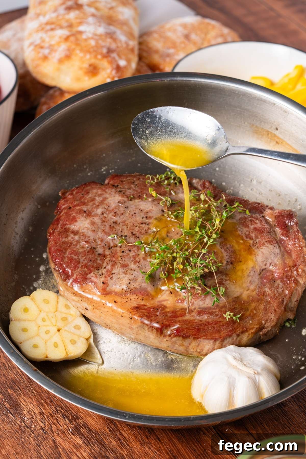 A spoon pours melted butter over a cooked steak with garlic and herbs in a pan.