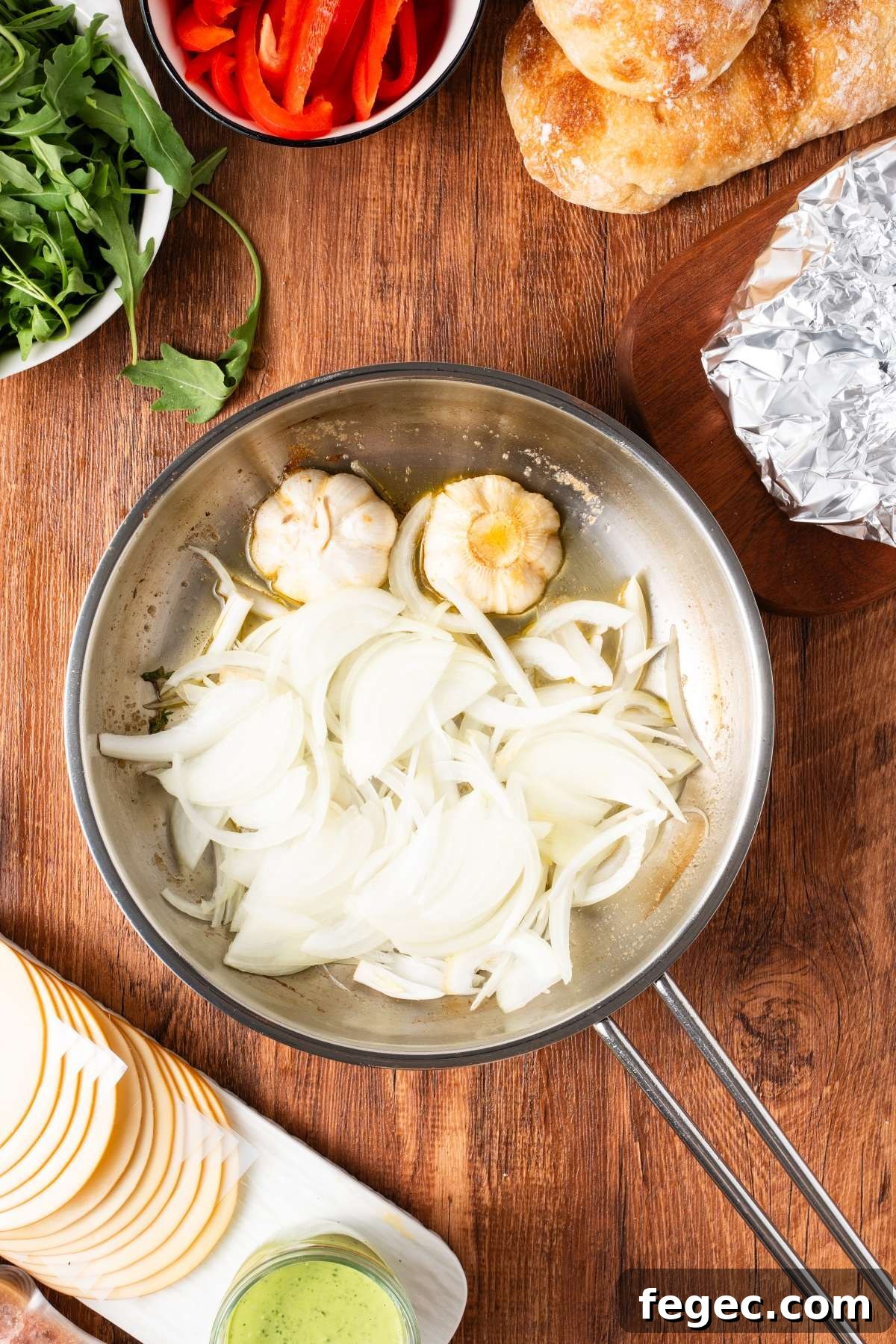 Sliced onions and halved garlic cooking in a skillet, surrounded by bread, cheese, greens, and red peppers.