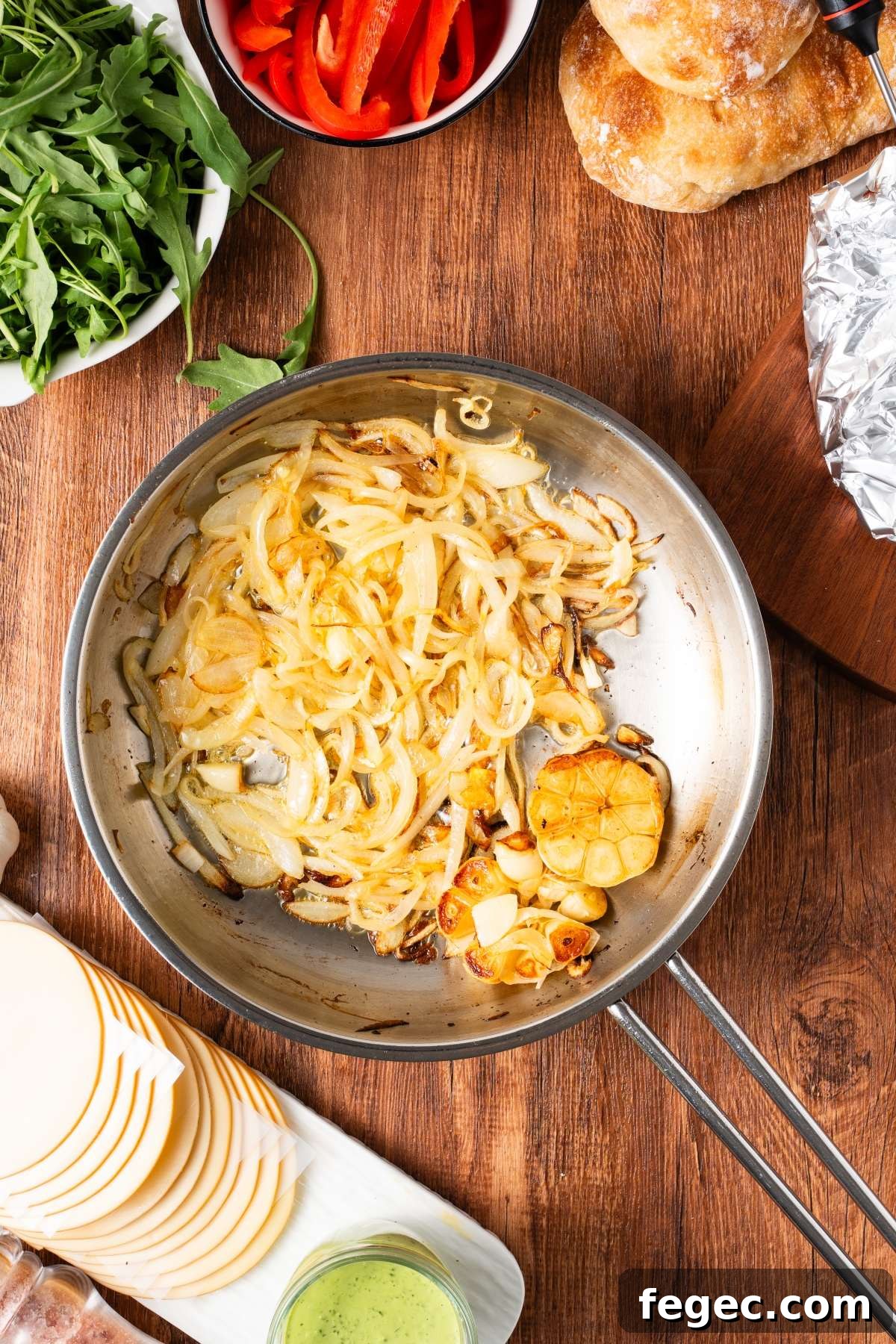 Sliced onions and garlic sautéing in a pan, surrounded by arugula, bread, red peppers, cheese, and sauce.