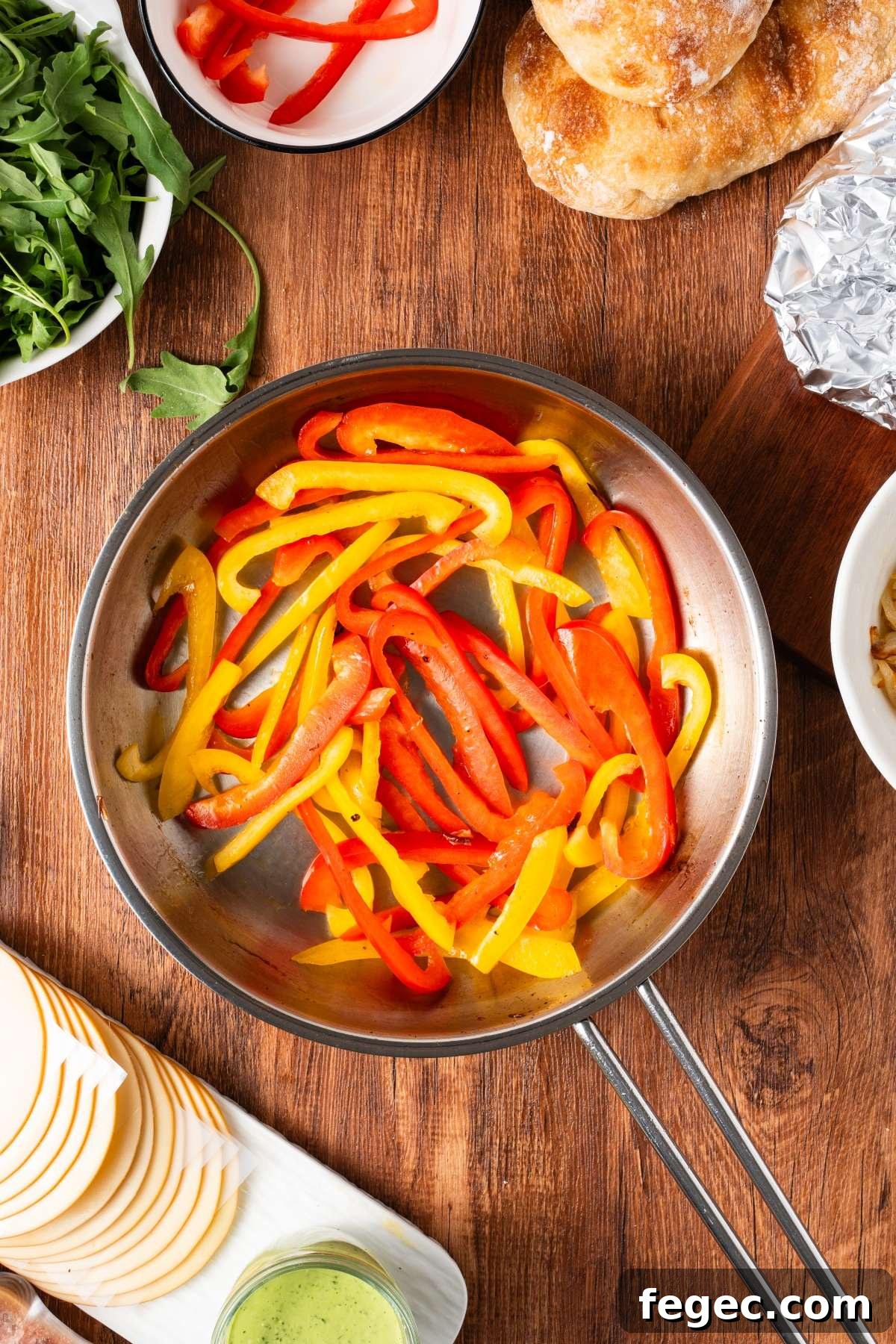 Red and yellow bell pepper strips cooking in a skillet, surrounded by sandwich ingredients on a wooden table.