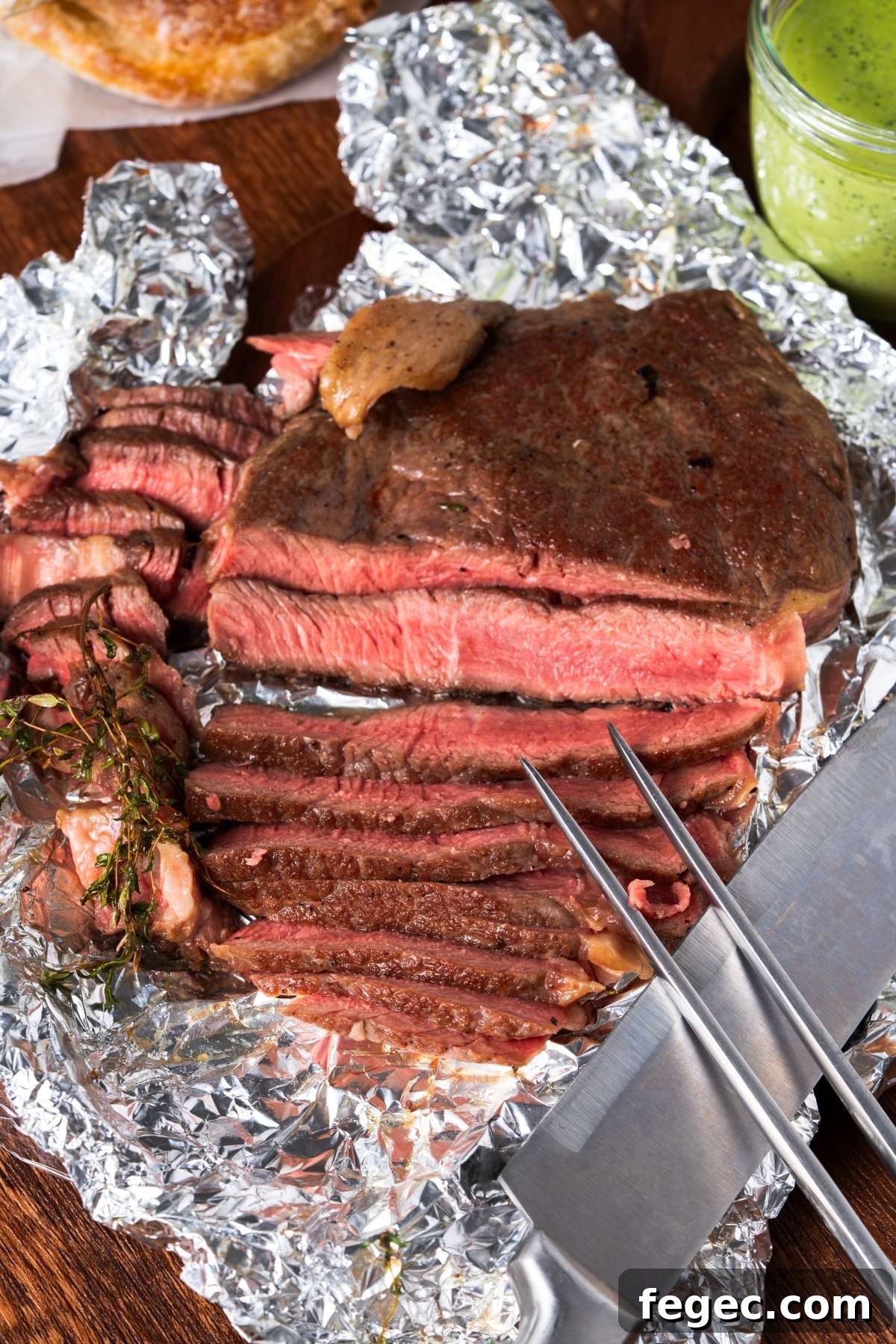 Sliced medium-rare steak on foil with a knife and fork, garnished with herbs.