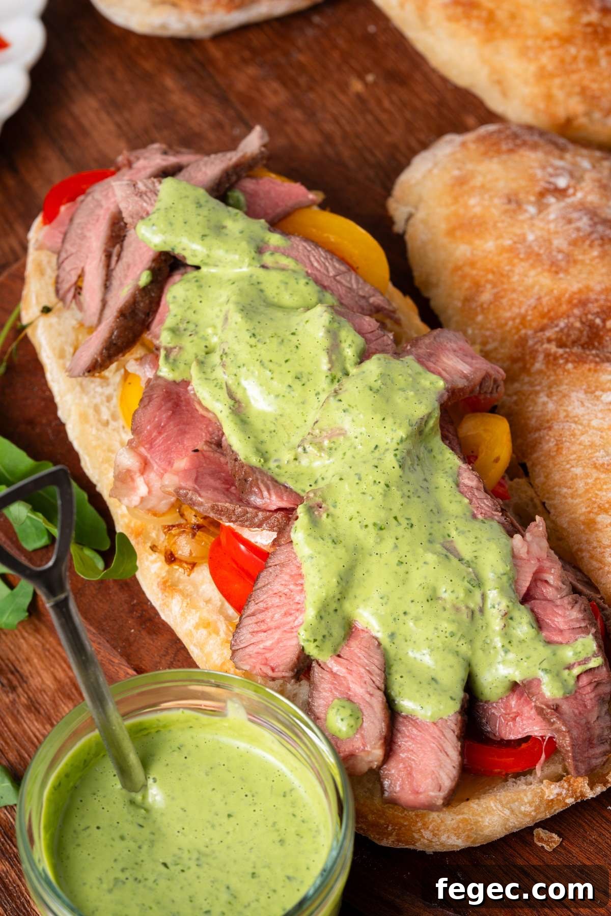 Sliced steak on ciabatta bread with yellow tomatoes and green sauce, served on a wooden board.