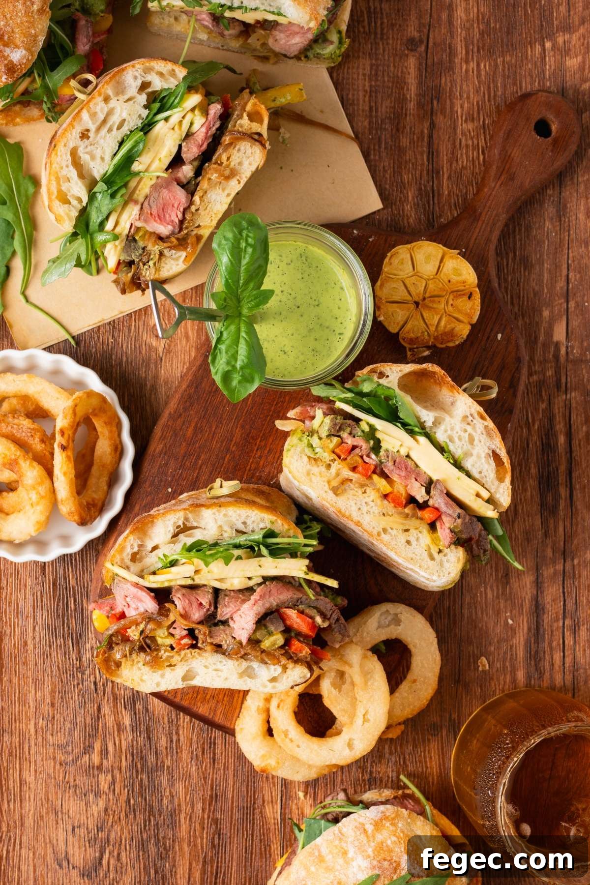 Sandwiches with steak, greens, cheese, and onion rings on a wooden table with green sauce and a drink.