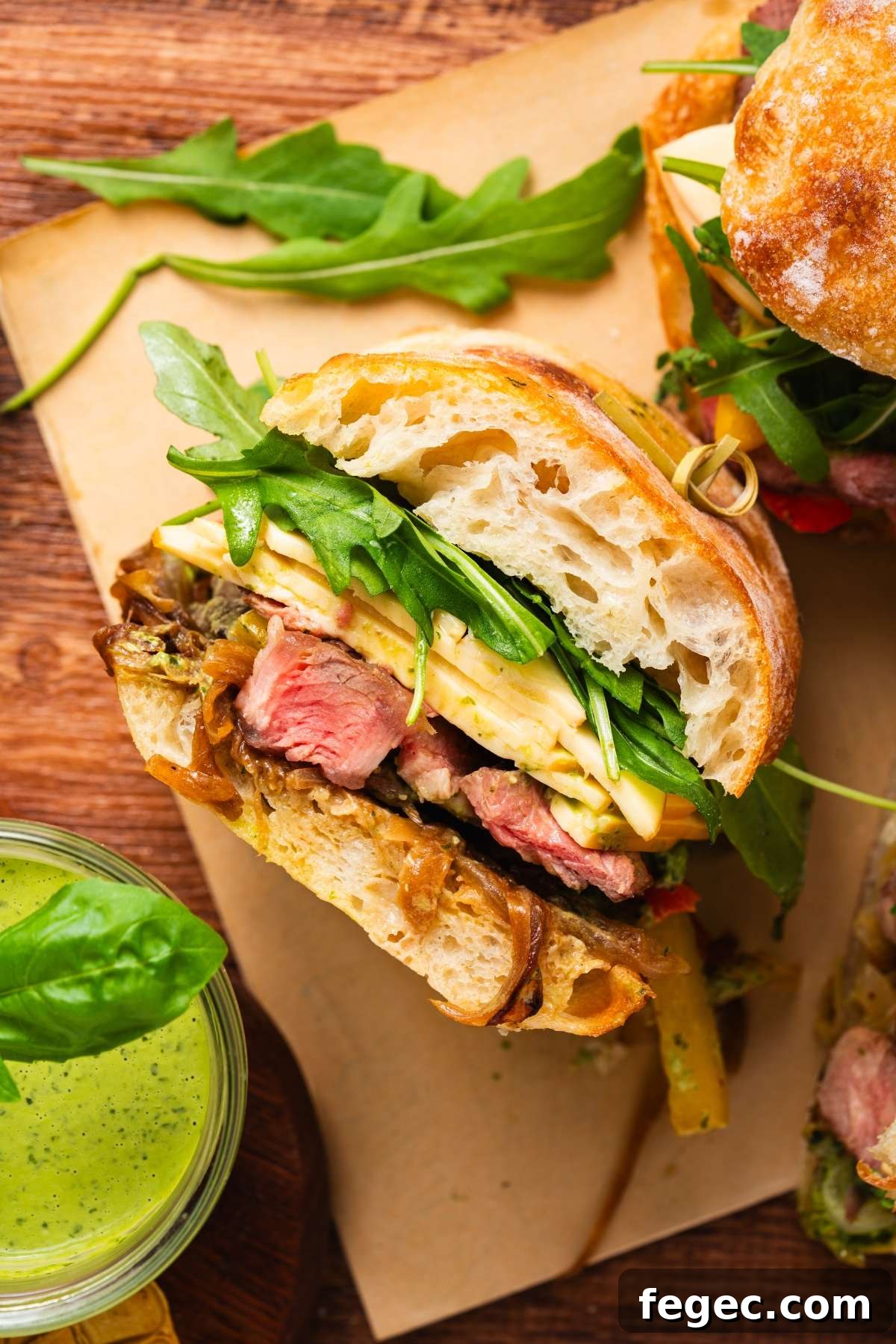 Ciabatta sandwich with steak, cheese, arugula, and grilled veggies, served beside a jar of green sauce.