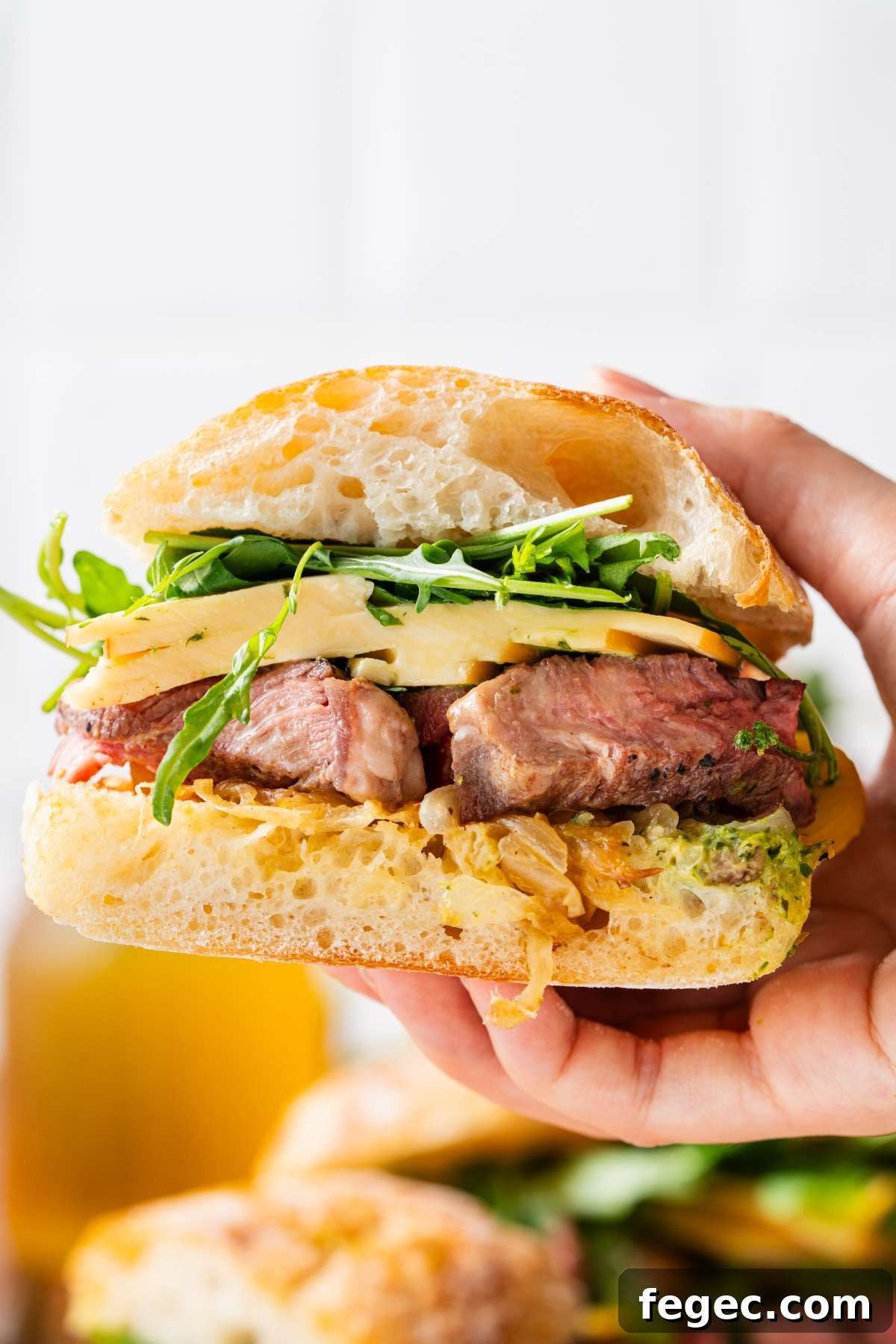 A hand holds a sandwich with steak, cheese, arugula, and sauce on ciabatta bread.