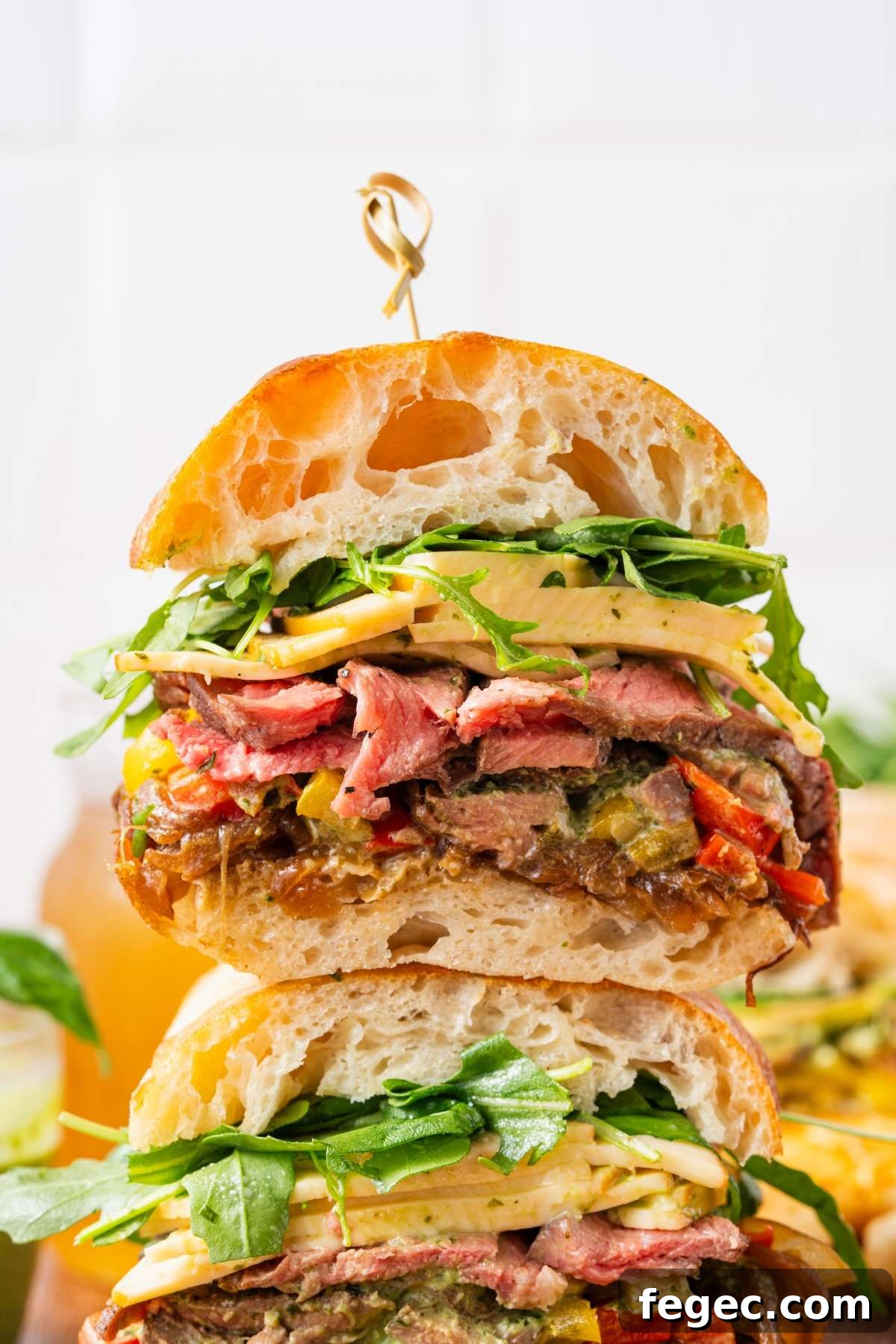 A stacked steak sandwich with cheese, arugula, and vegetables on rustic bread, secured with a skewer.